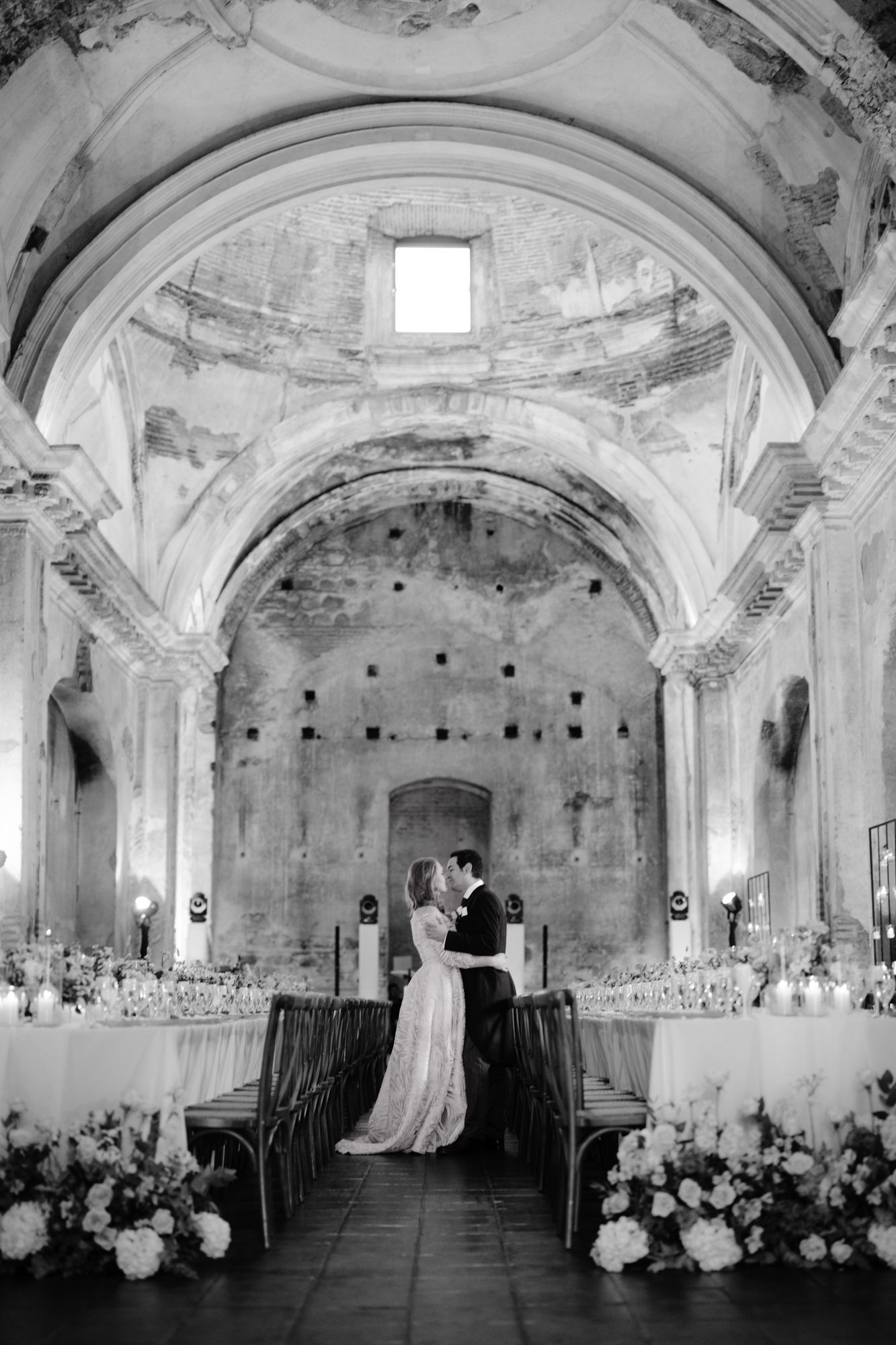 A black and white wedding portrait showing a couple embracing in an elegant historic venue.