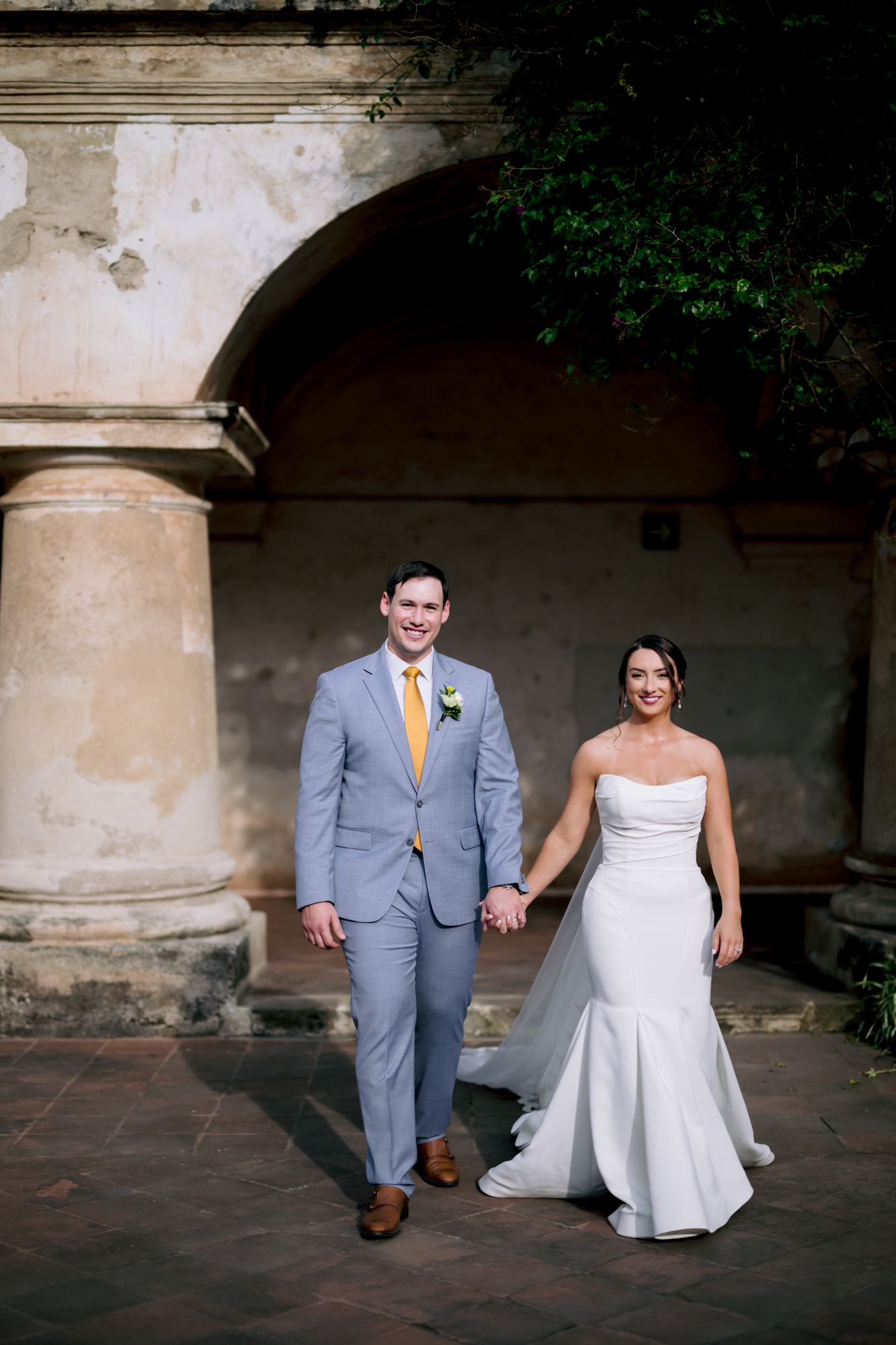 Stylish couple in wedding attire walks through historic stone courtyard with yellow accents.