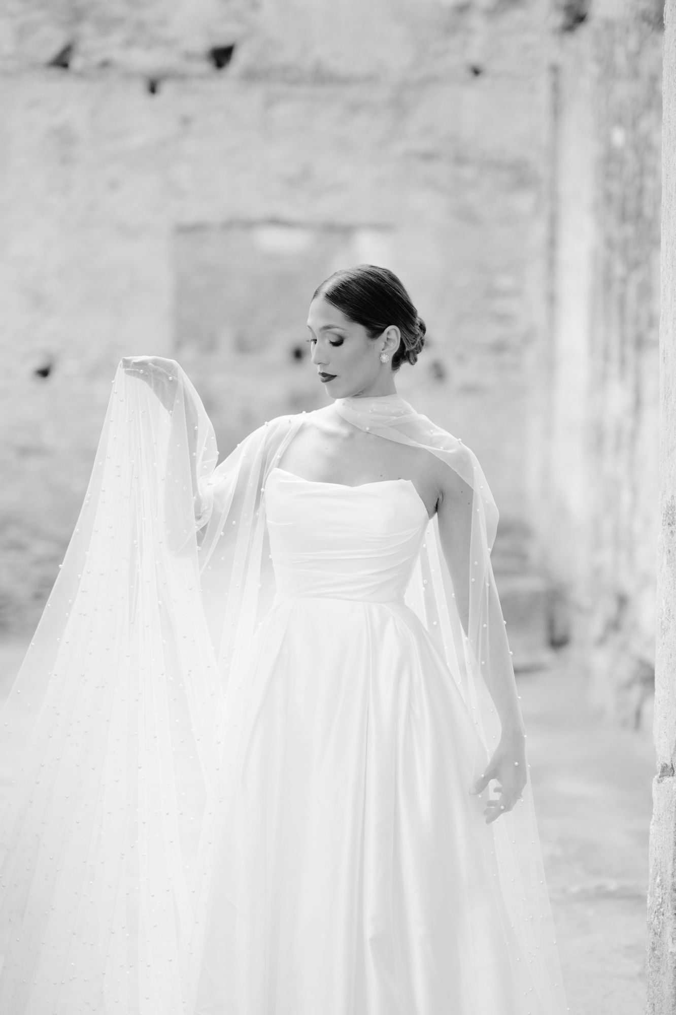 Black and white portrait of bride in strapless gown holding flowing veil against stone wall.