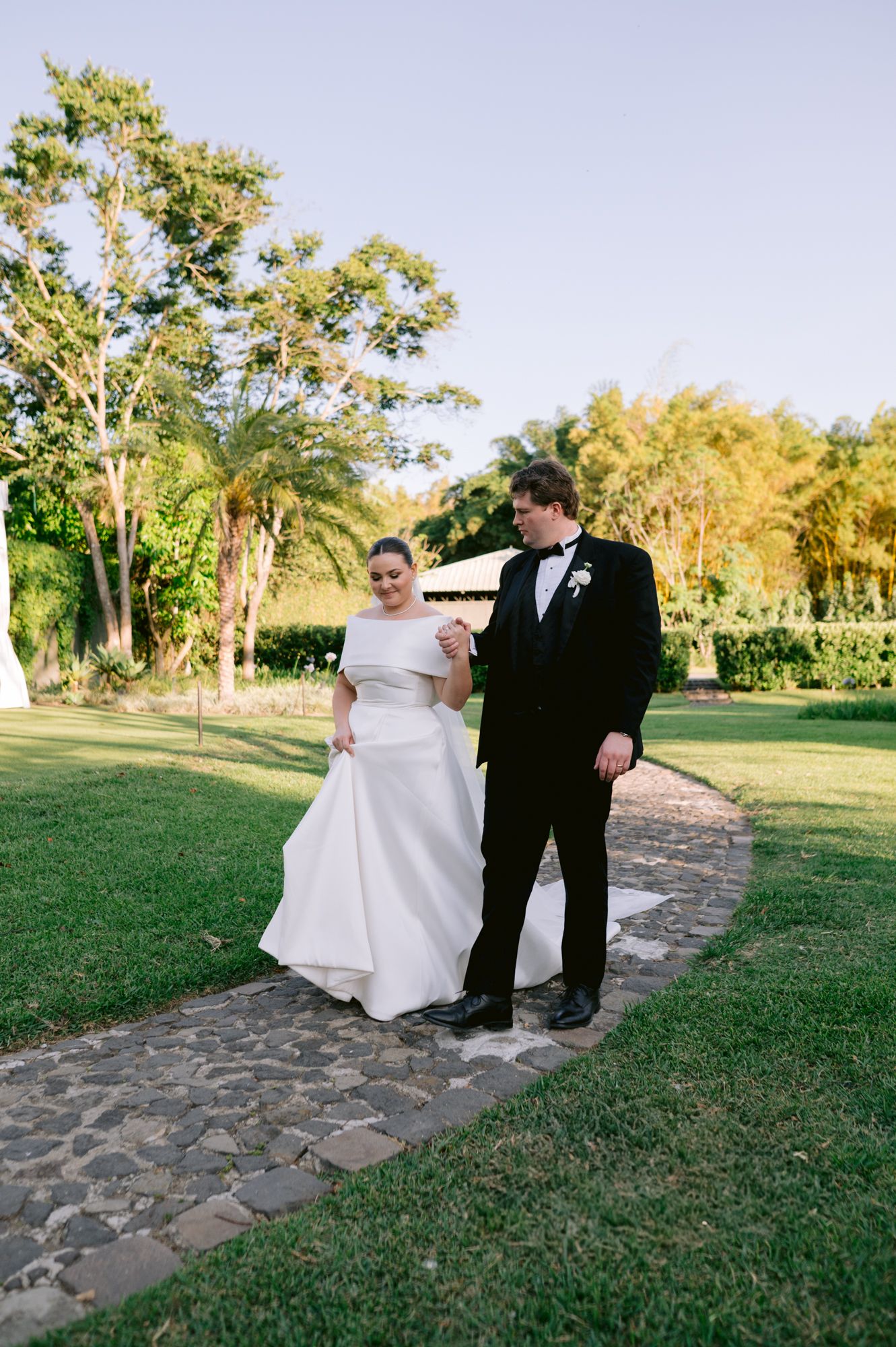 Couple in wedding attire walking along a stone path through a manicured garden landscape.