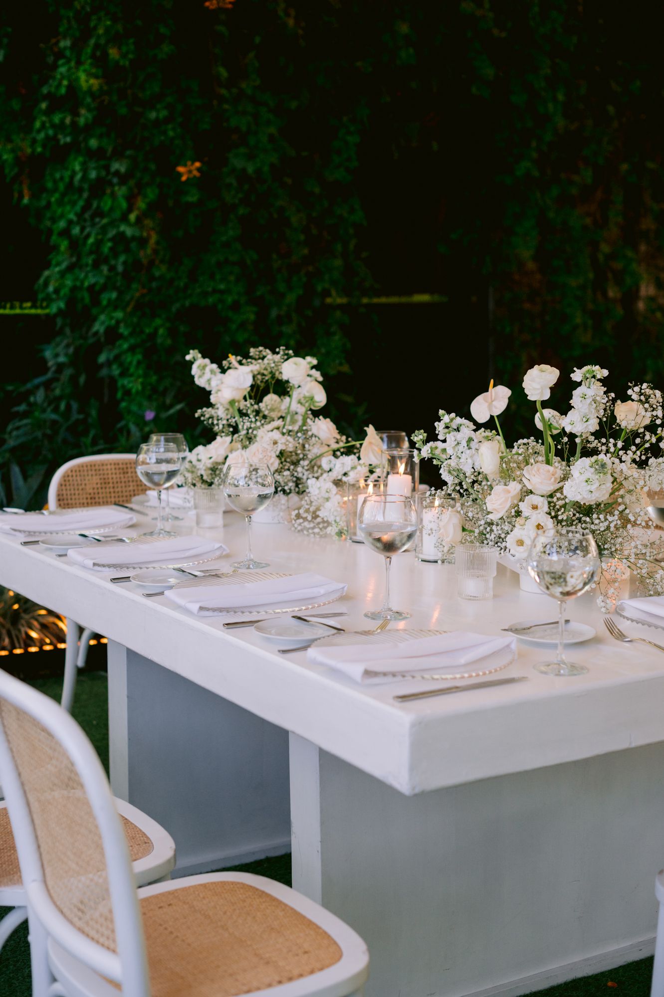 Elegant white wedding table setting with delicate floral centerpieces and candlelight against a dark green backdrop.