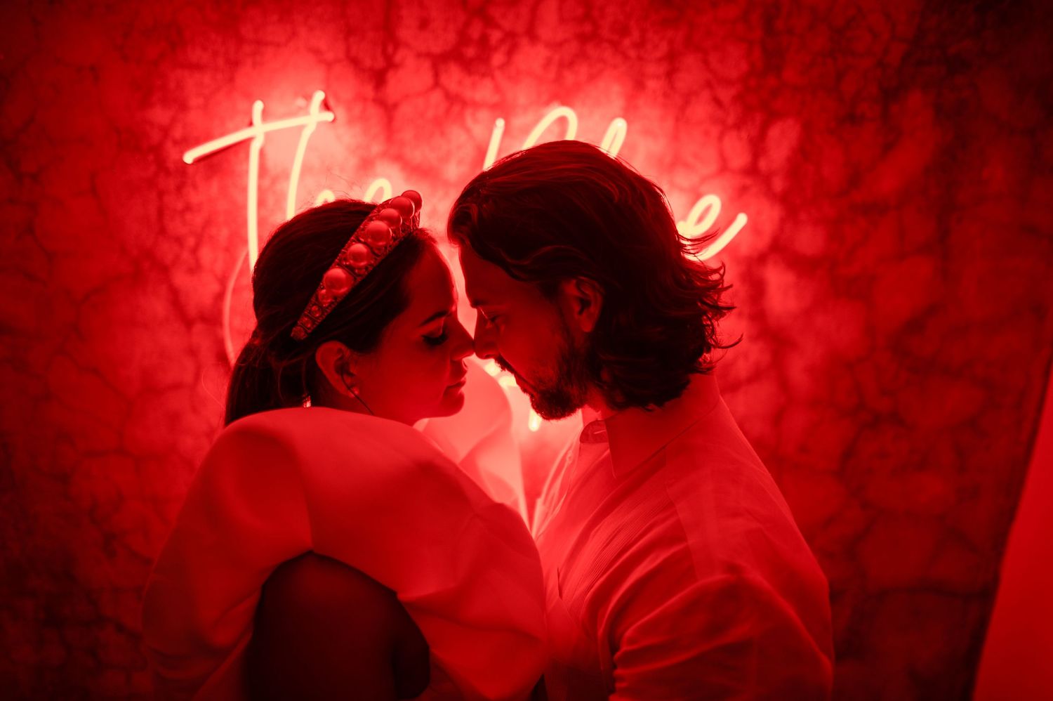 A romantic couple shares an intimate moment under dramatic red lighting with a neon sign in the background.