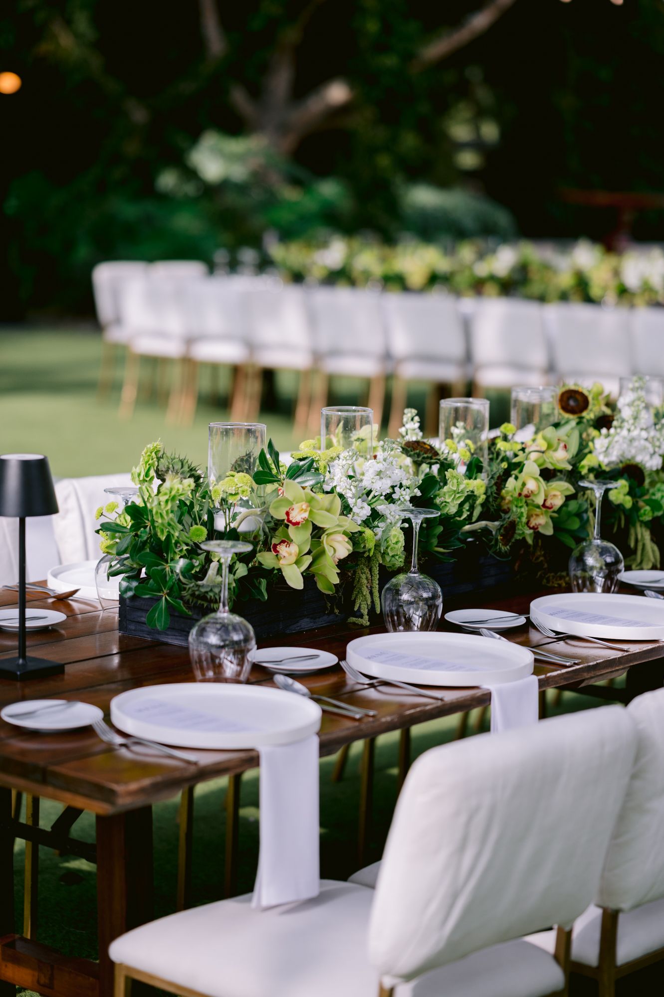 Elegant outdoor wedding table setting with white chairs, wooden tables and lush green floral centerpieces.