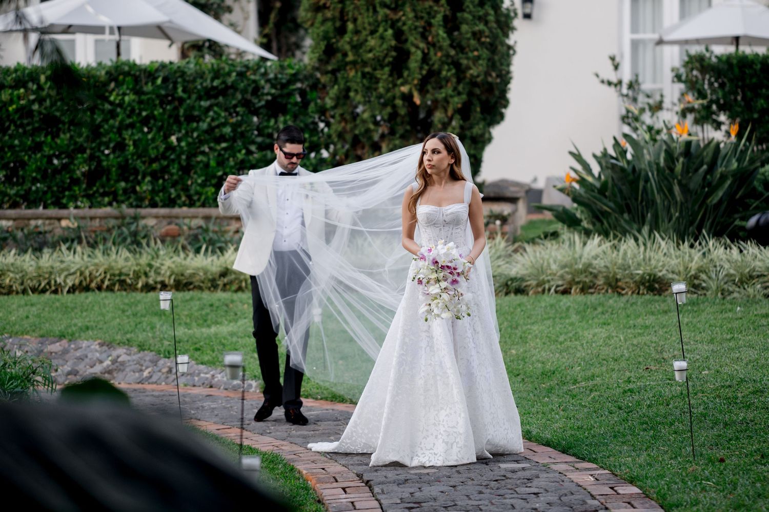 Elegant wedding couple posing on garden path as groom helps adjust bride's flowing veil in outdoor setting.