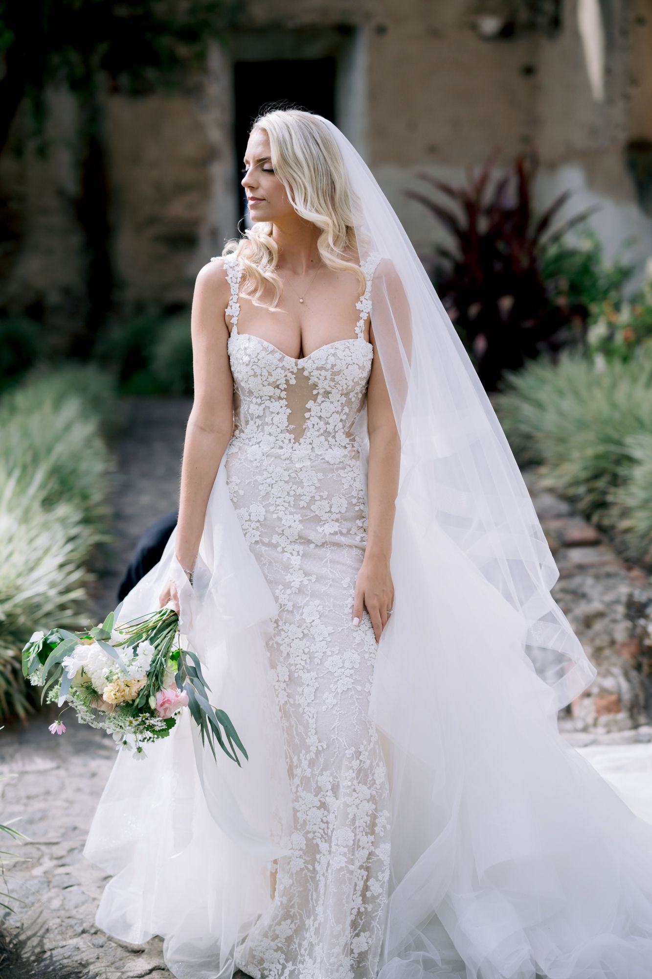 Bride in form-fitting lace gown holds bouquet against stone wall backdrop.