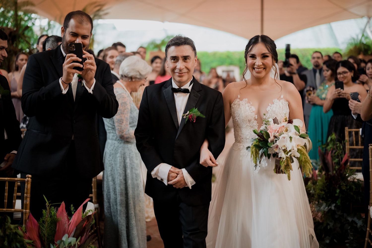 A wedding ceremony procession in an outdoor venue under a peach-colored tent.