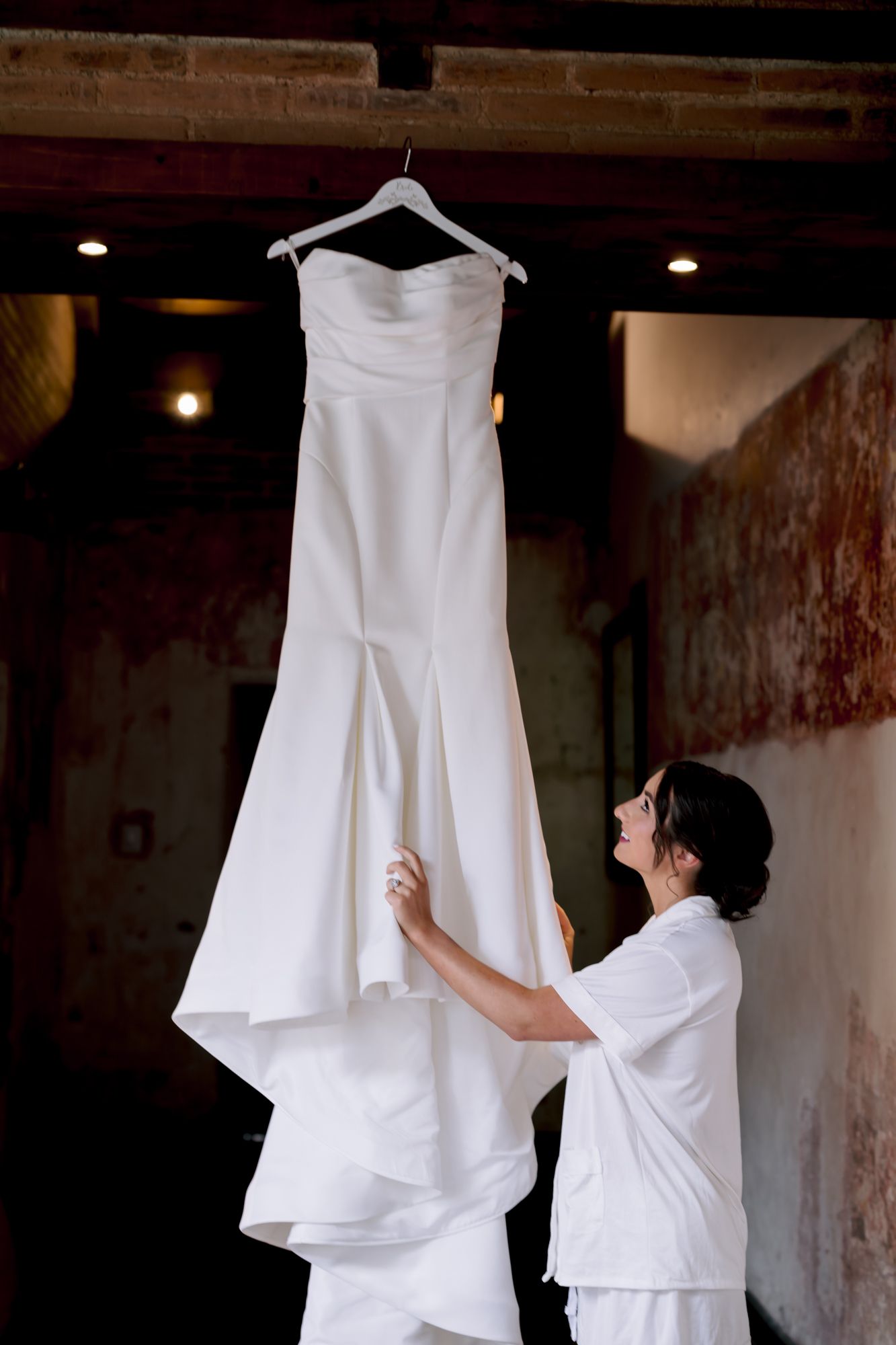 White strapless wedding dress hanging against rustic wall while being prepared.