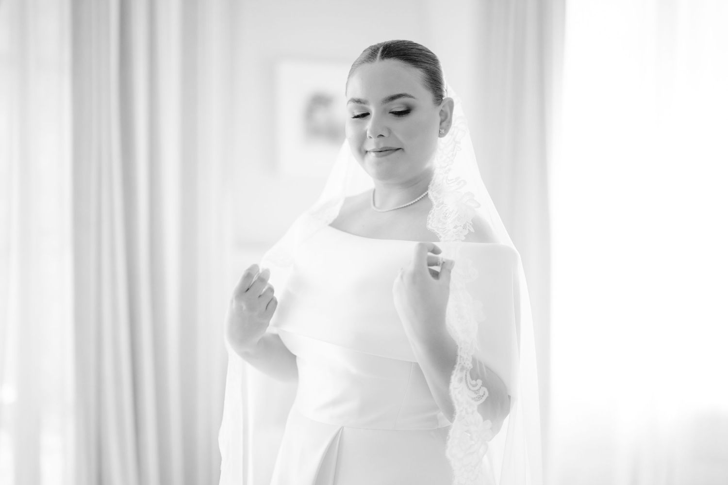 Black and white portrait of bride adjusting veil in natural window light.