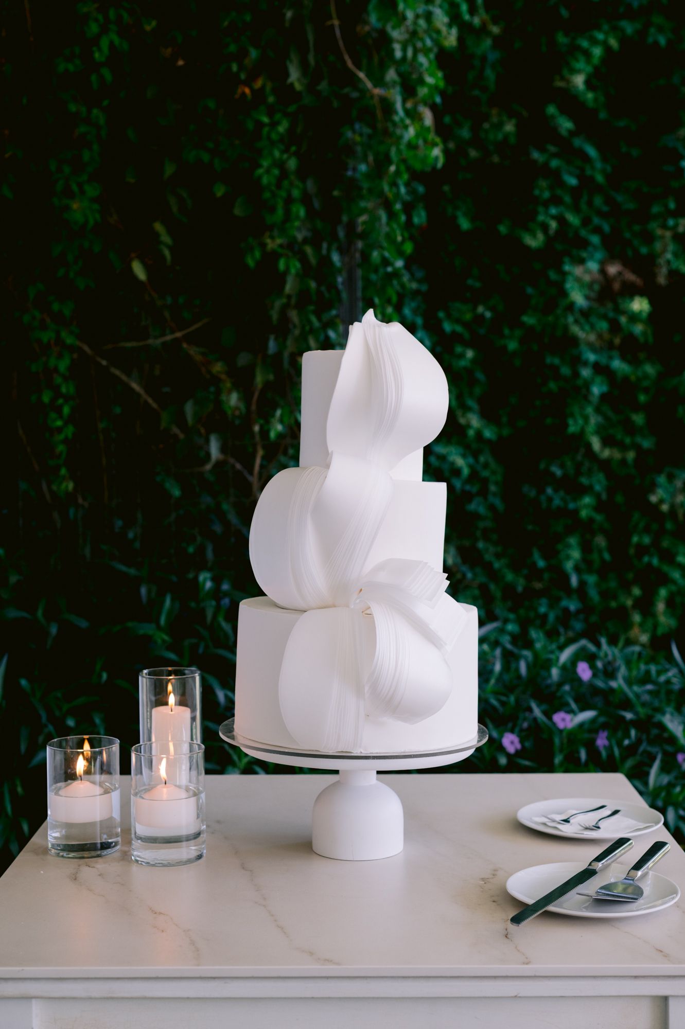 Modern white sculptural wedding cake with geometric design elements displayed with candles against dark foliage.