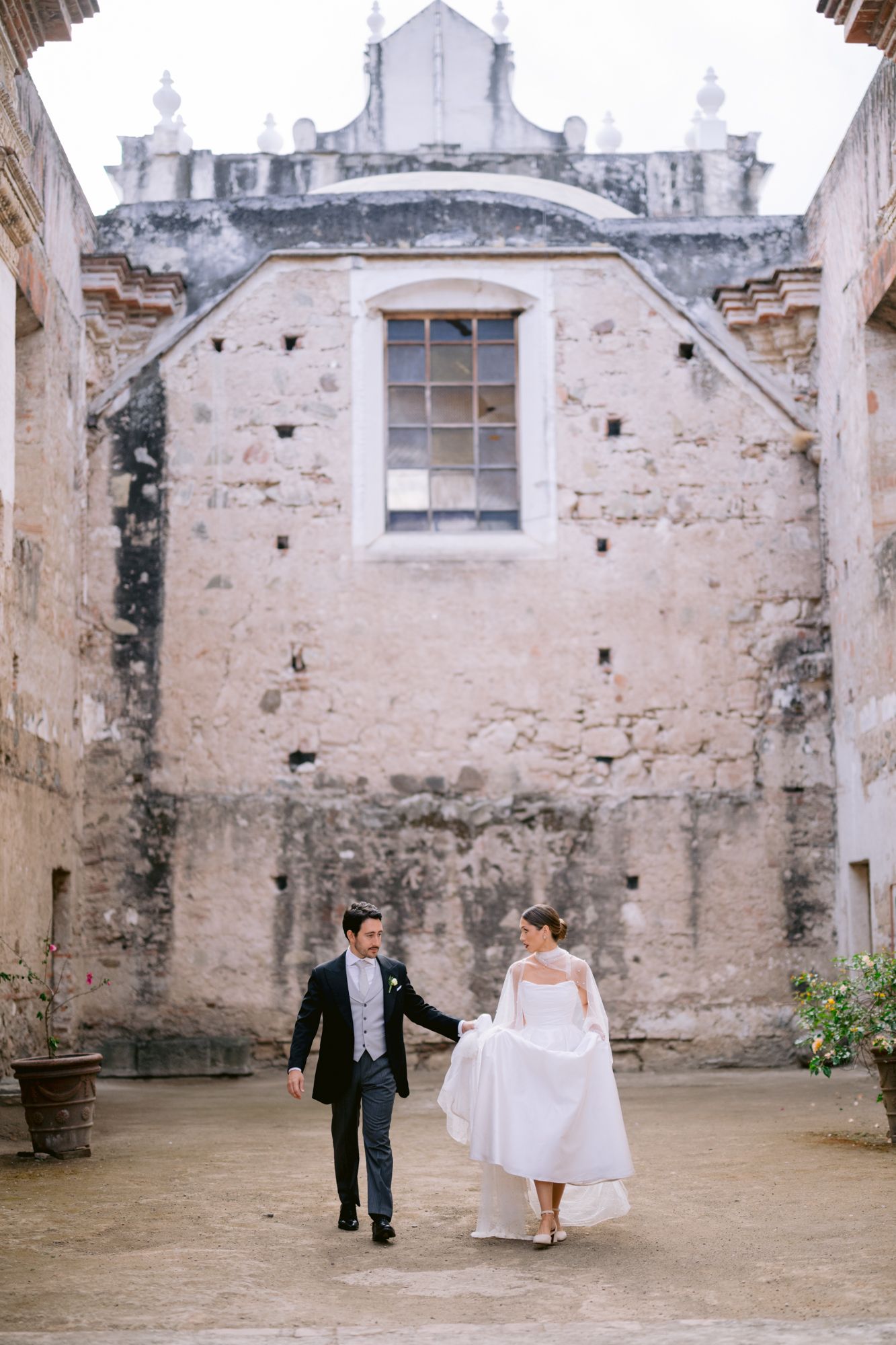 Wedding couple walking hand in hand through rustic stone courtyard with weathered walls.