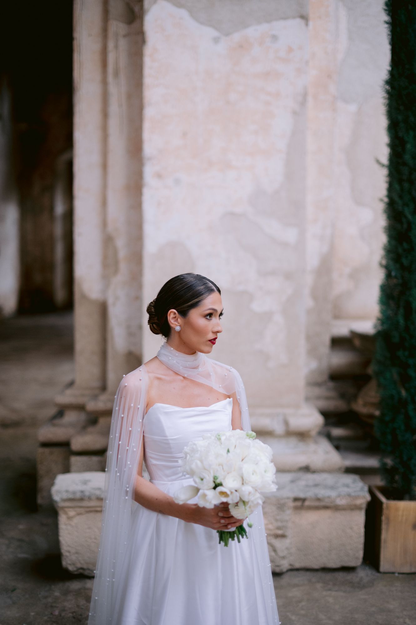 Bride in elegant white gown with cape holding bouquet of white flowers against stone wall.