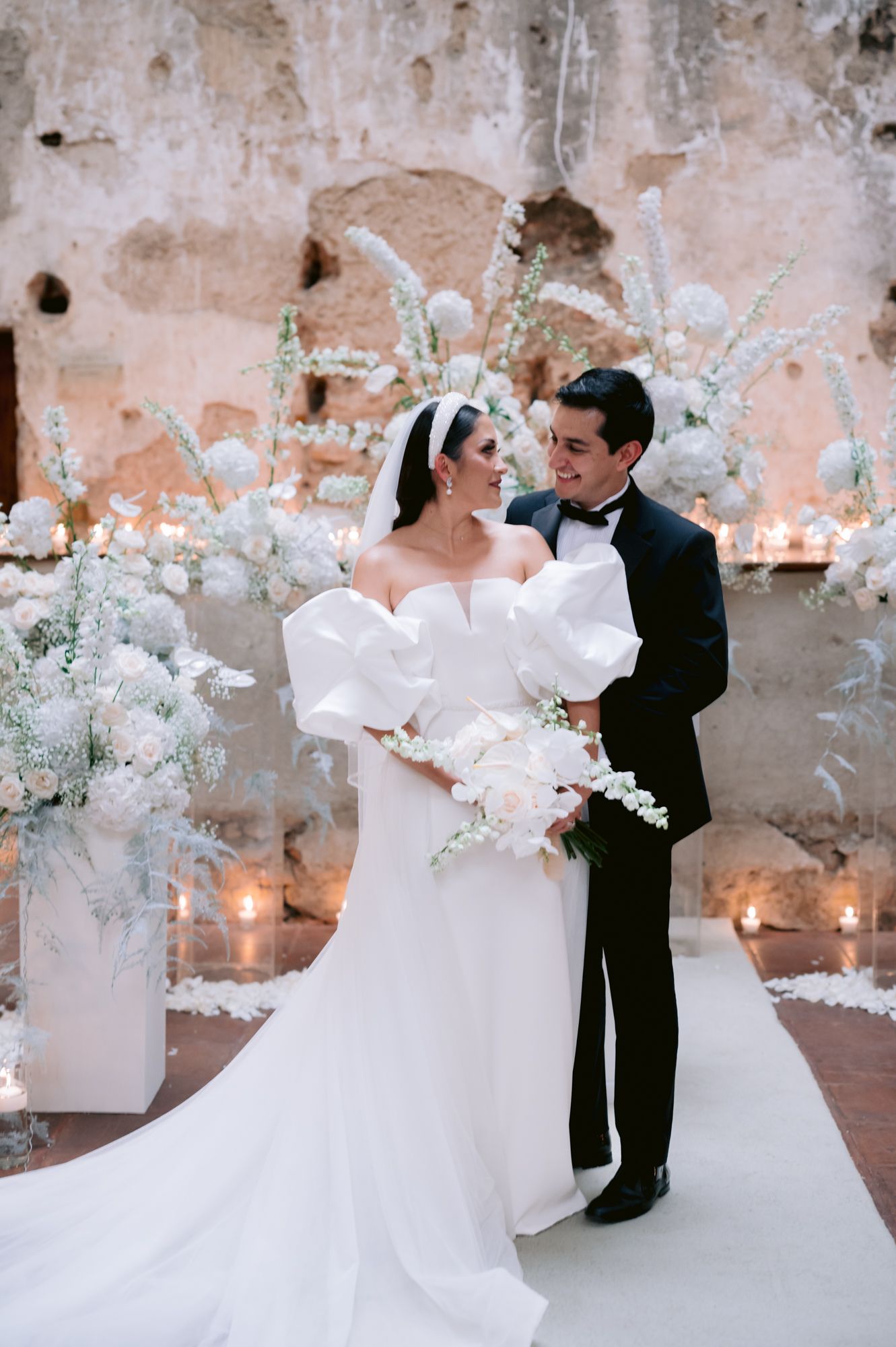 Elegant wedding couple poses together against a textured wall with white floral arrangements and candlelight.