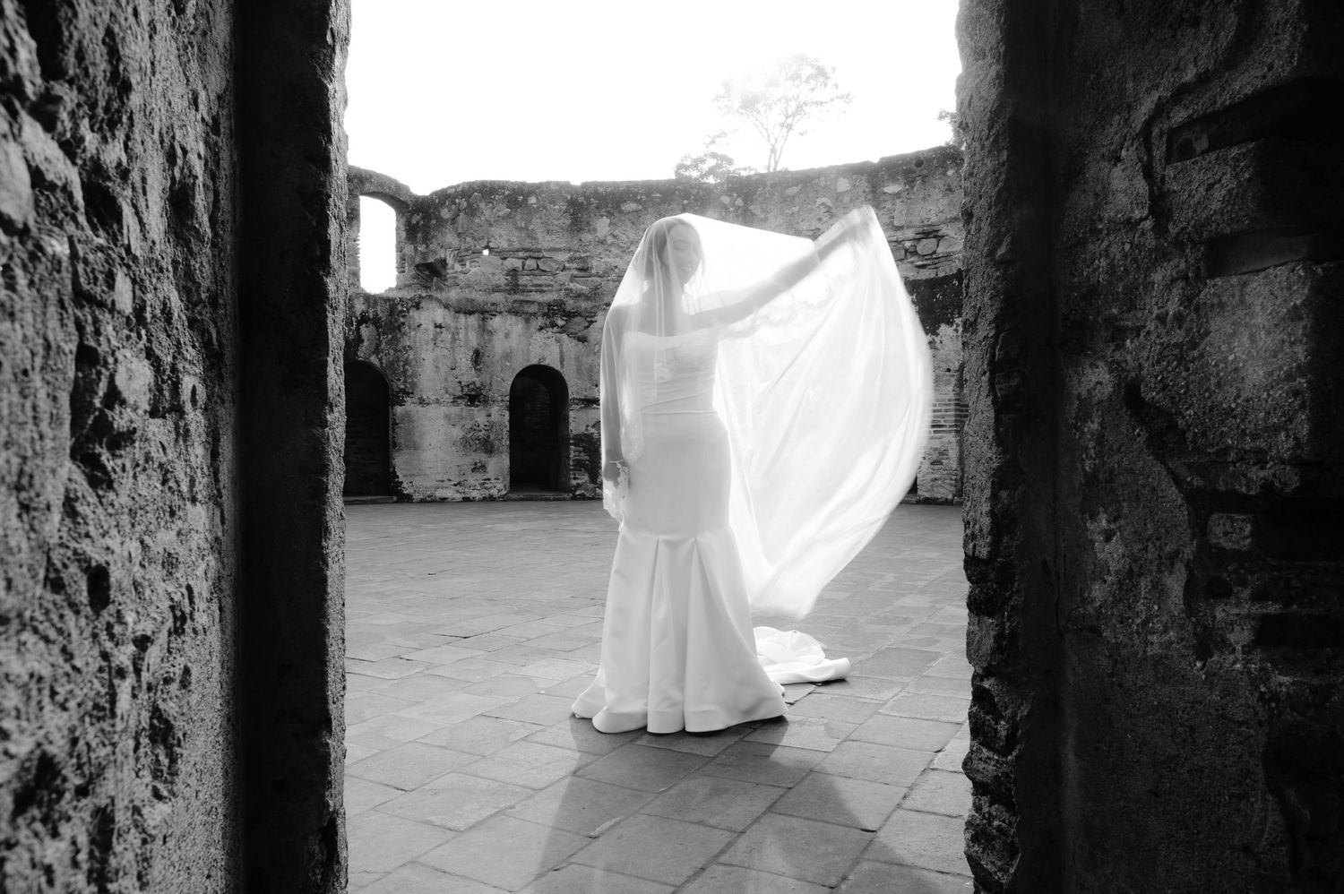 Black and white bridal portrait showing flowing dress and veil in historic stone courtyard.