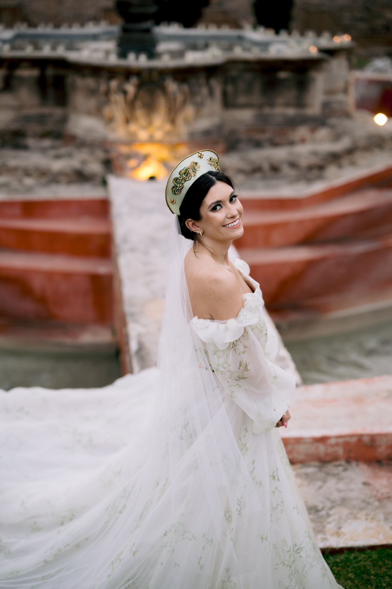 Elegant wedding dress with off-shoulder design and flowing veil against rustic stone backdrop.