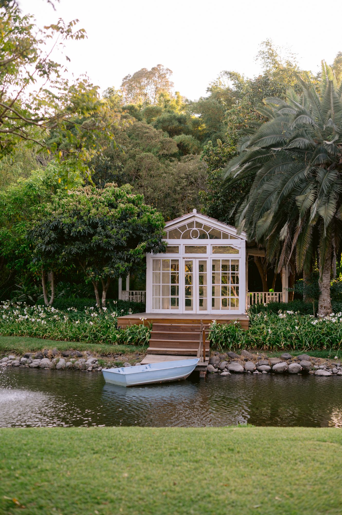 White gazebo overlooking a pond with a small boat, surrounded by palm trees and lush garden landscaping.