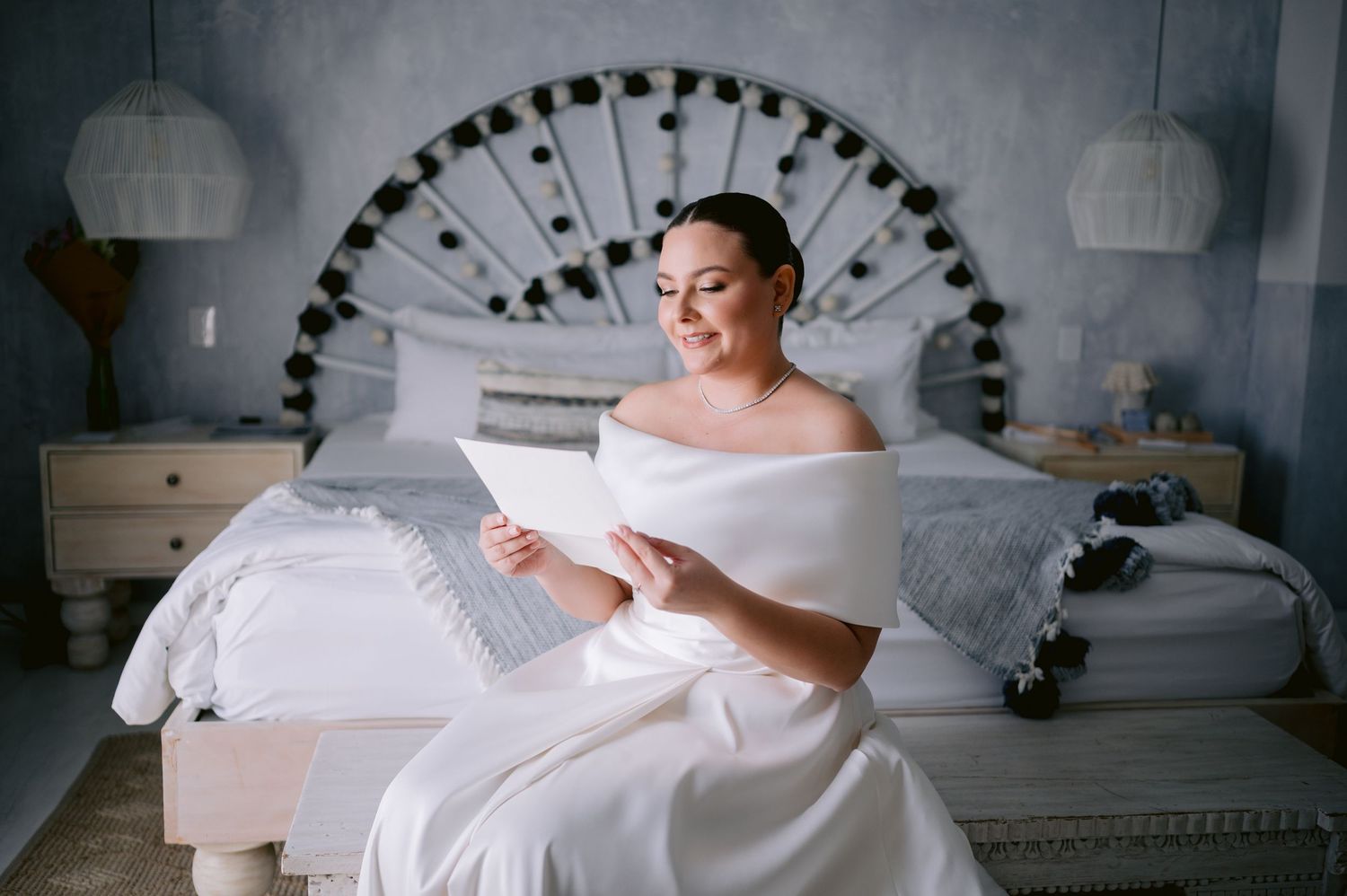 Bride in white off-shoulder gown reading while seated on bed with decorative headboard.
