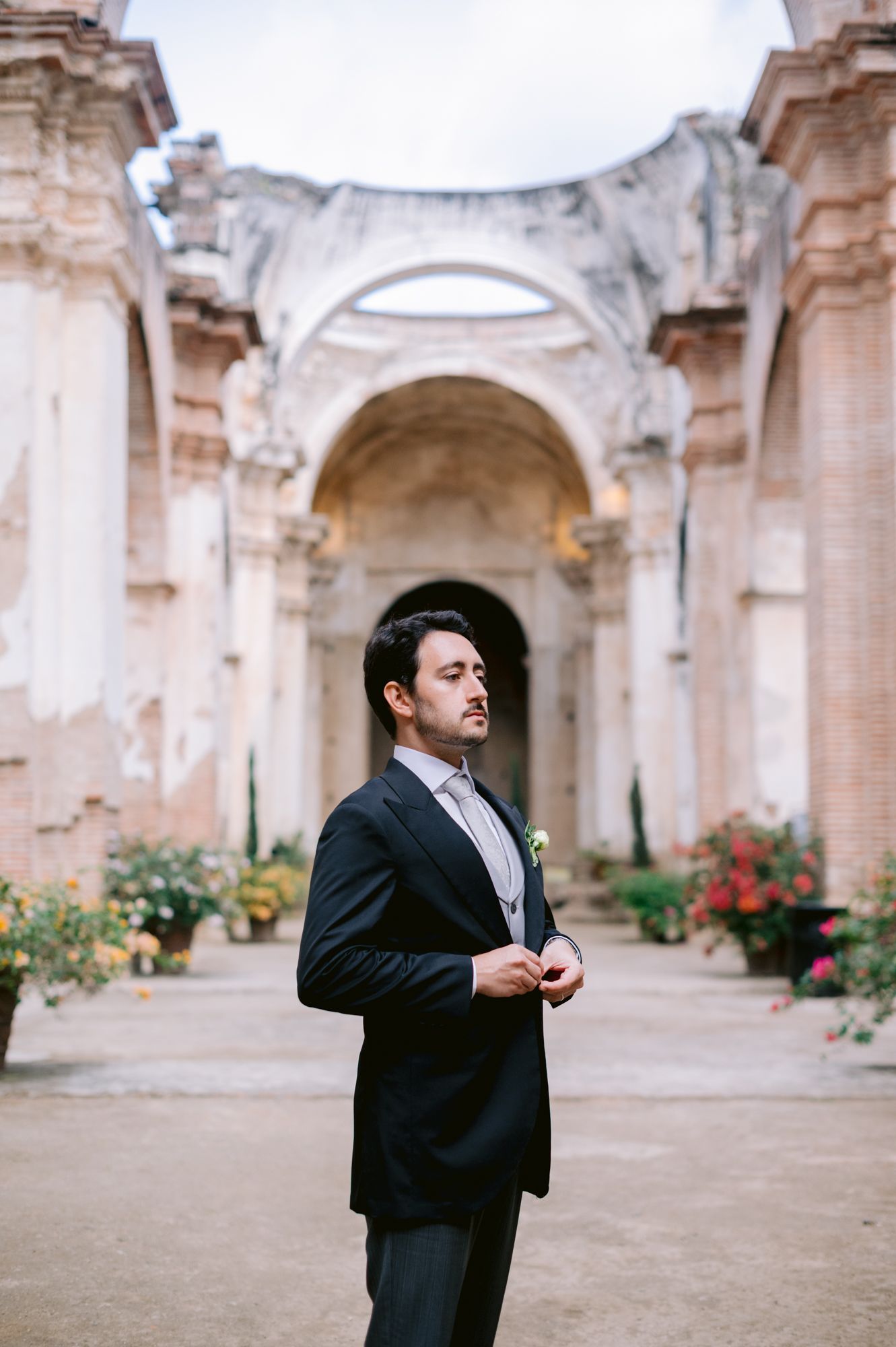 Groom in classic black tuxedo standing in ornate stone courtyard with arched architecture.