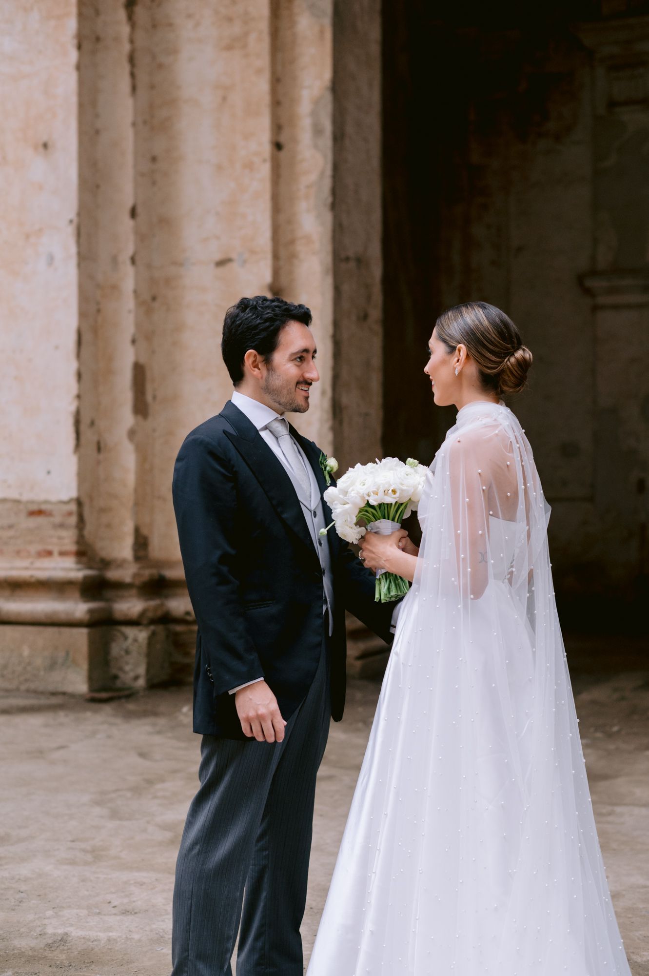 Elegant couple in wedding attire share moment together against stone church backdrop.