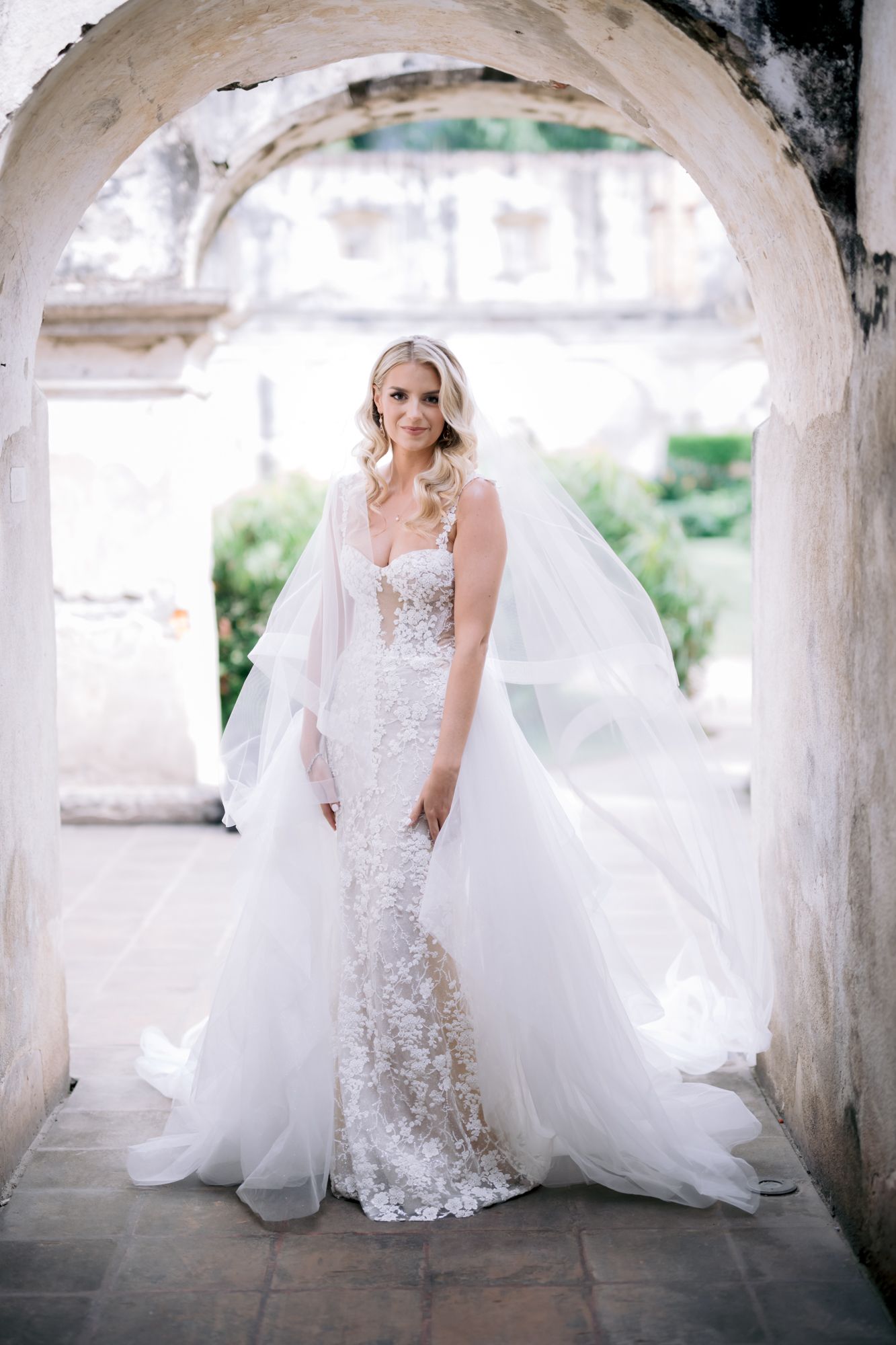 Bride in elegant lace wedding gown with flowing veil poses under rustic stone archway.