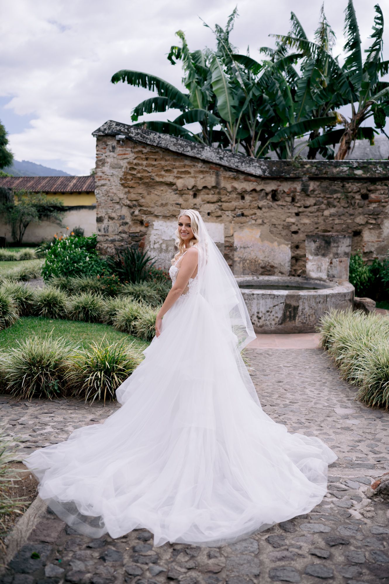 Bride in white wedding gown walks along stone path with tropical foliage and ruins in background.