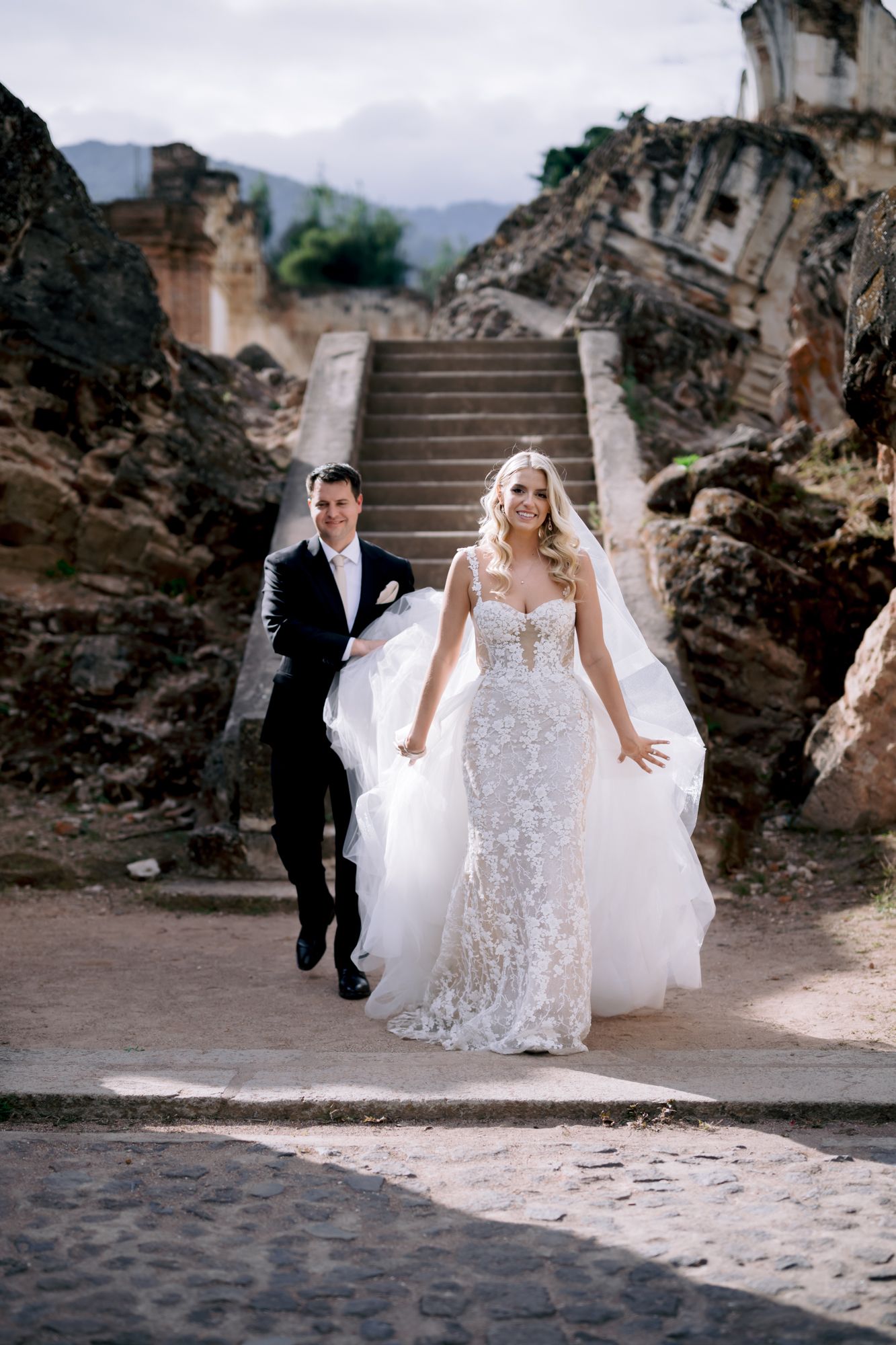 A wedding couple descends ornate stone steps at a historic ruins venue.