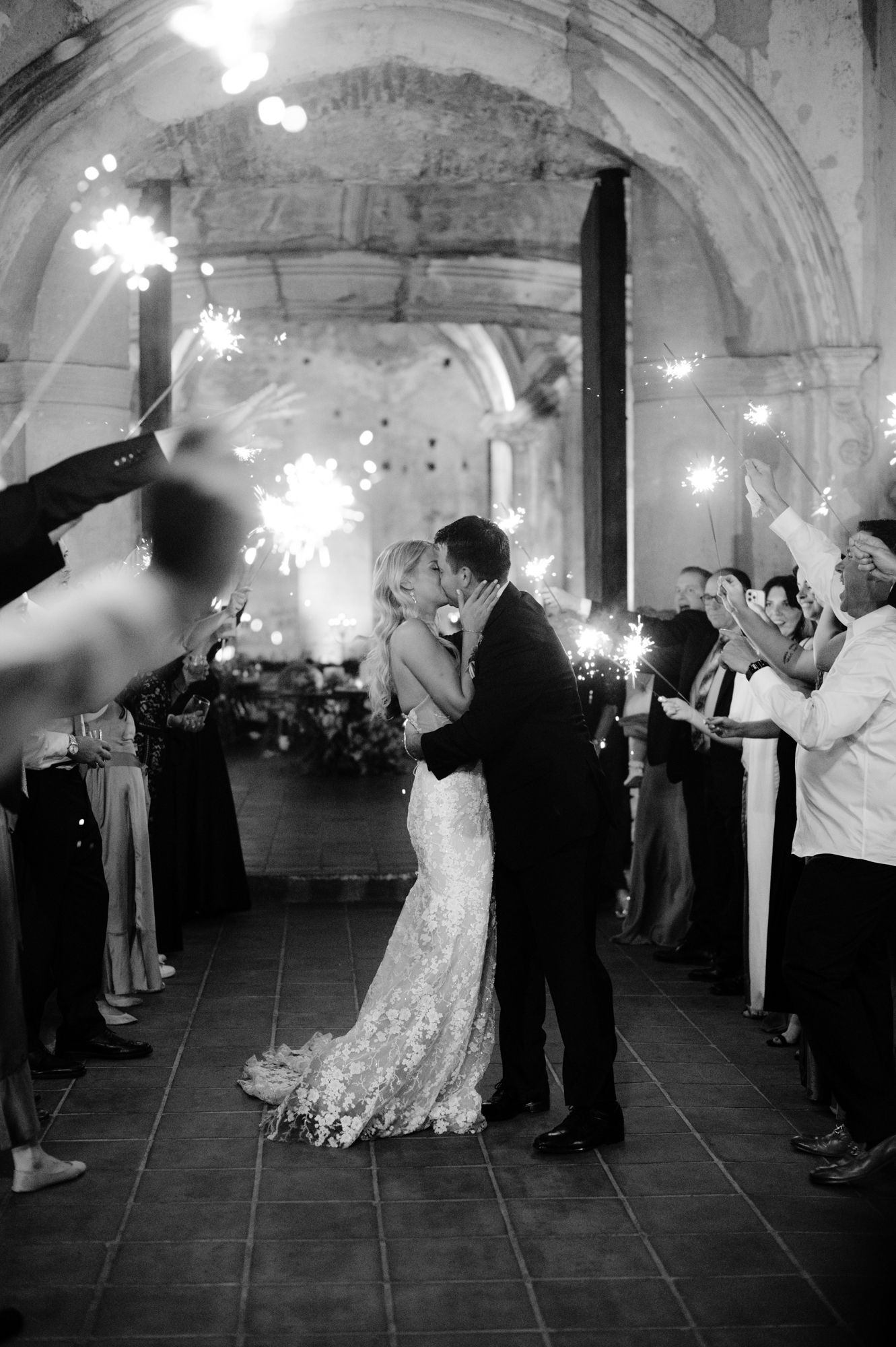 Newlyweds share romantic moment under sparklers held by wedding guests in historic venue.
