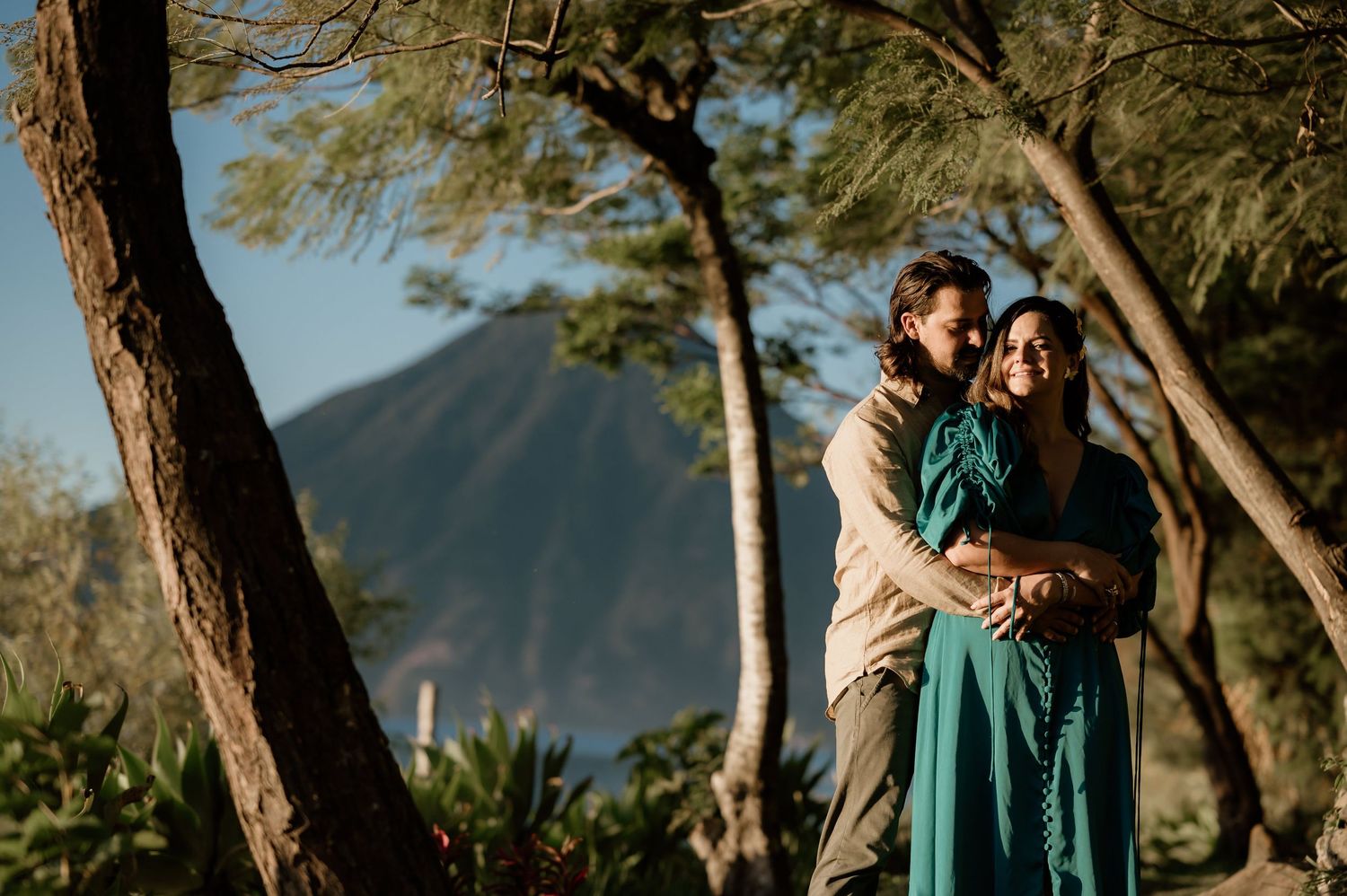 Couple embraces during sunset engagement photoshoot in a scenic mountain landscape with trees.
