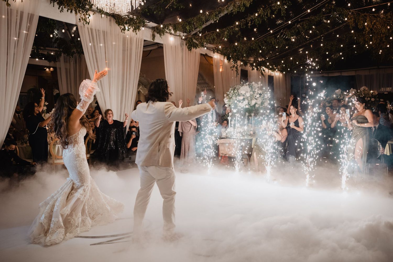 Wedding couple dancing amid sparklers and fog effects under string lights at their reception.
