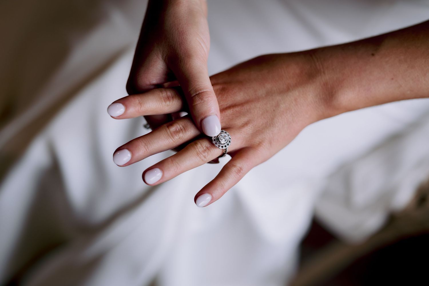 Close-up of hands showing off an engagement ring against white fabric.