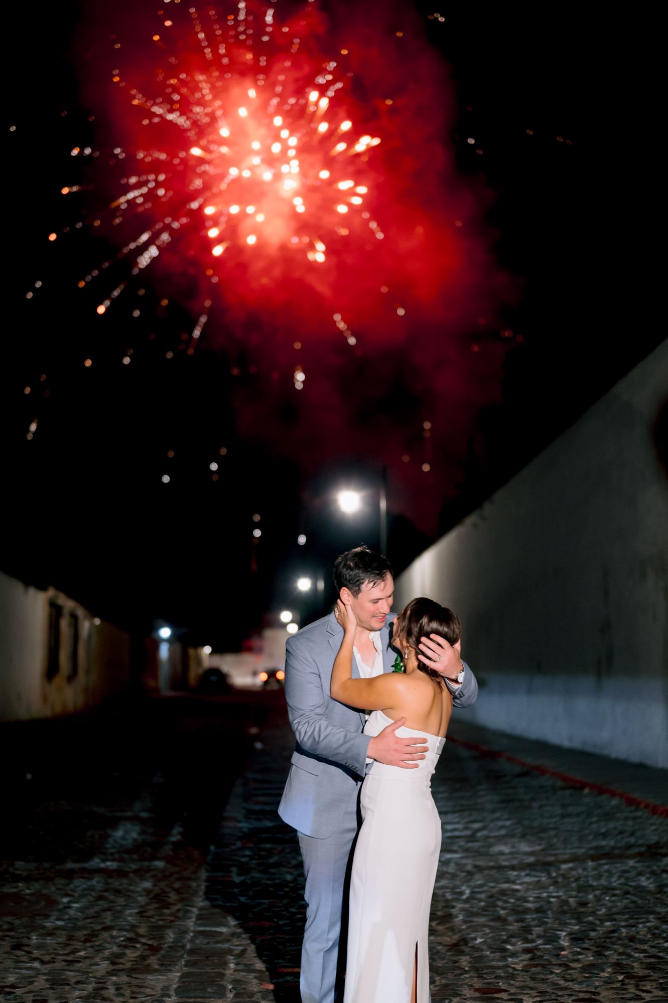 A couple embraces under bright red fireworks in the night sky.