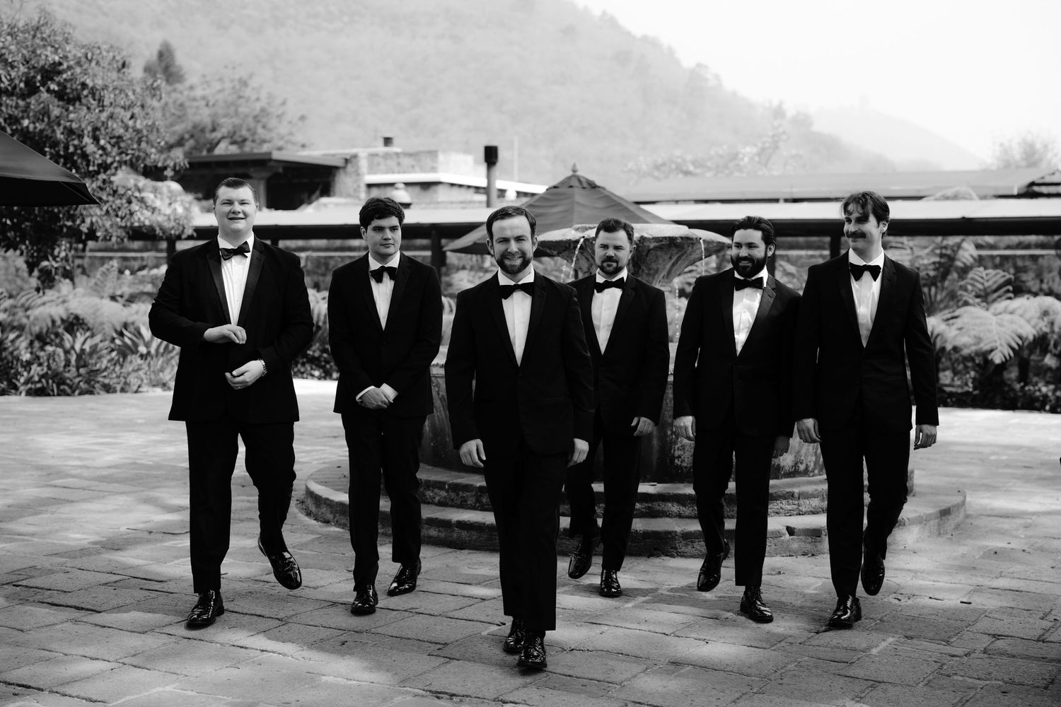 Group of groomsmen in black tuxedos walking together outdoors in classic black and white photography.