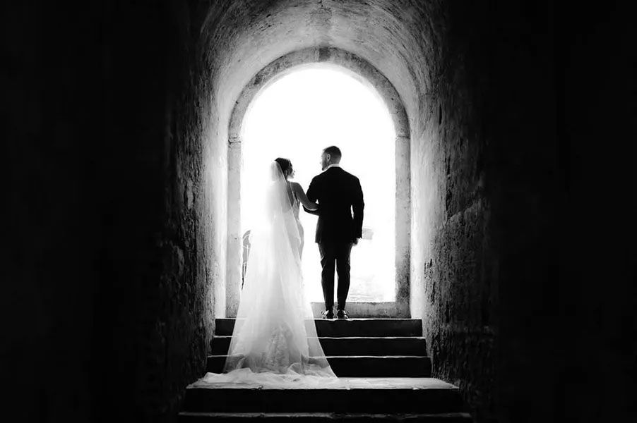 Silhouetted couple in wedding attire walking up stone steps through an archway tunnel illuminated by natural light.