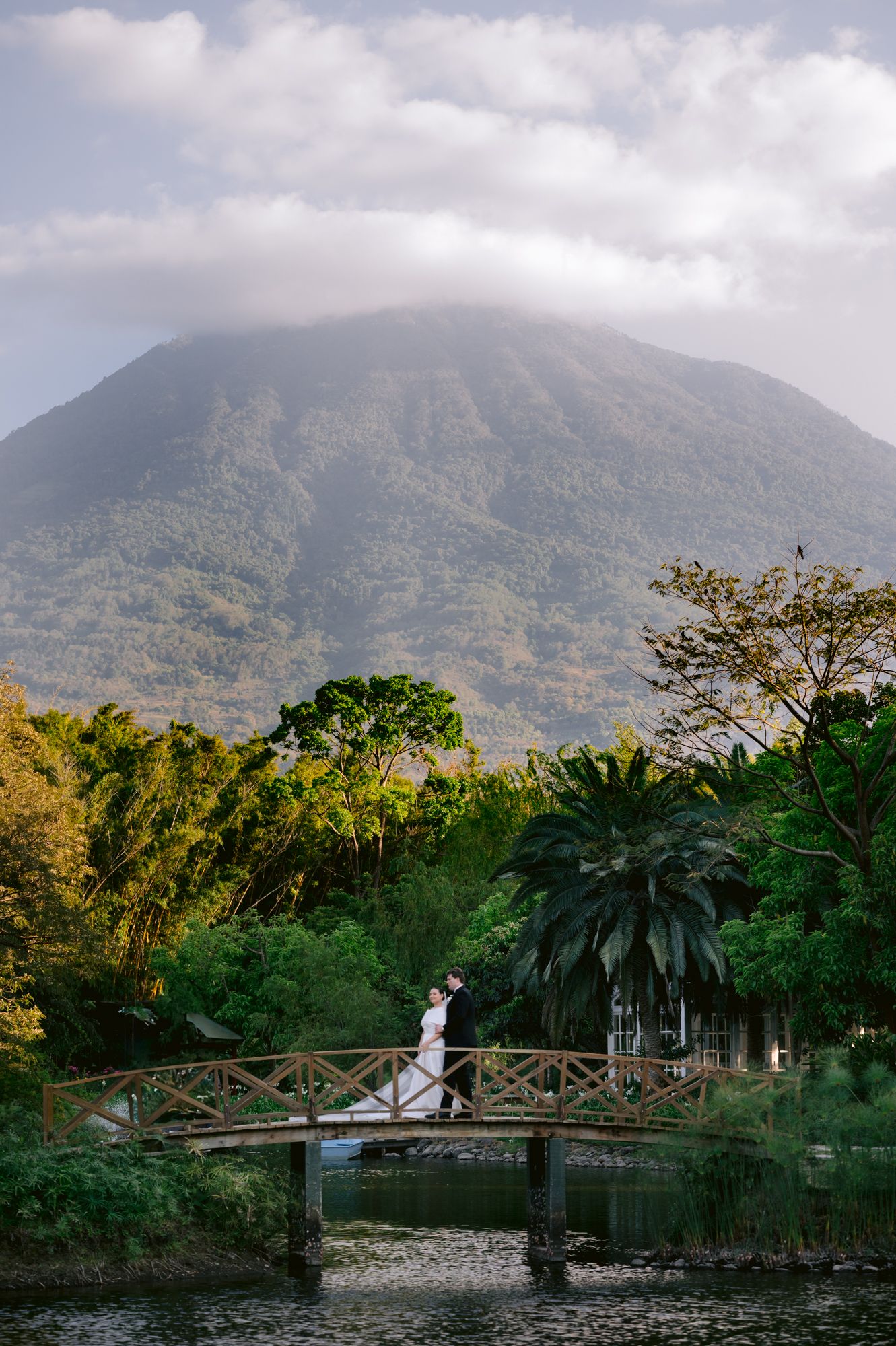 A wooden footbridge spans a pond with a majestic mountain rising in the background among lush tropical foliage.