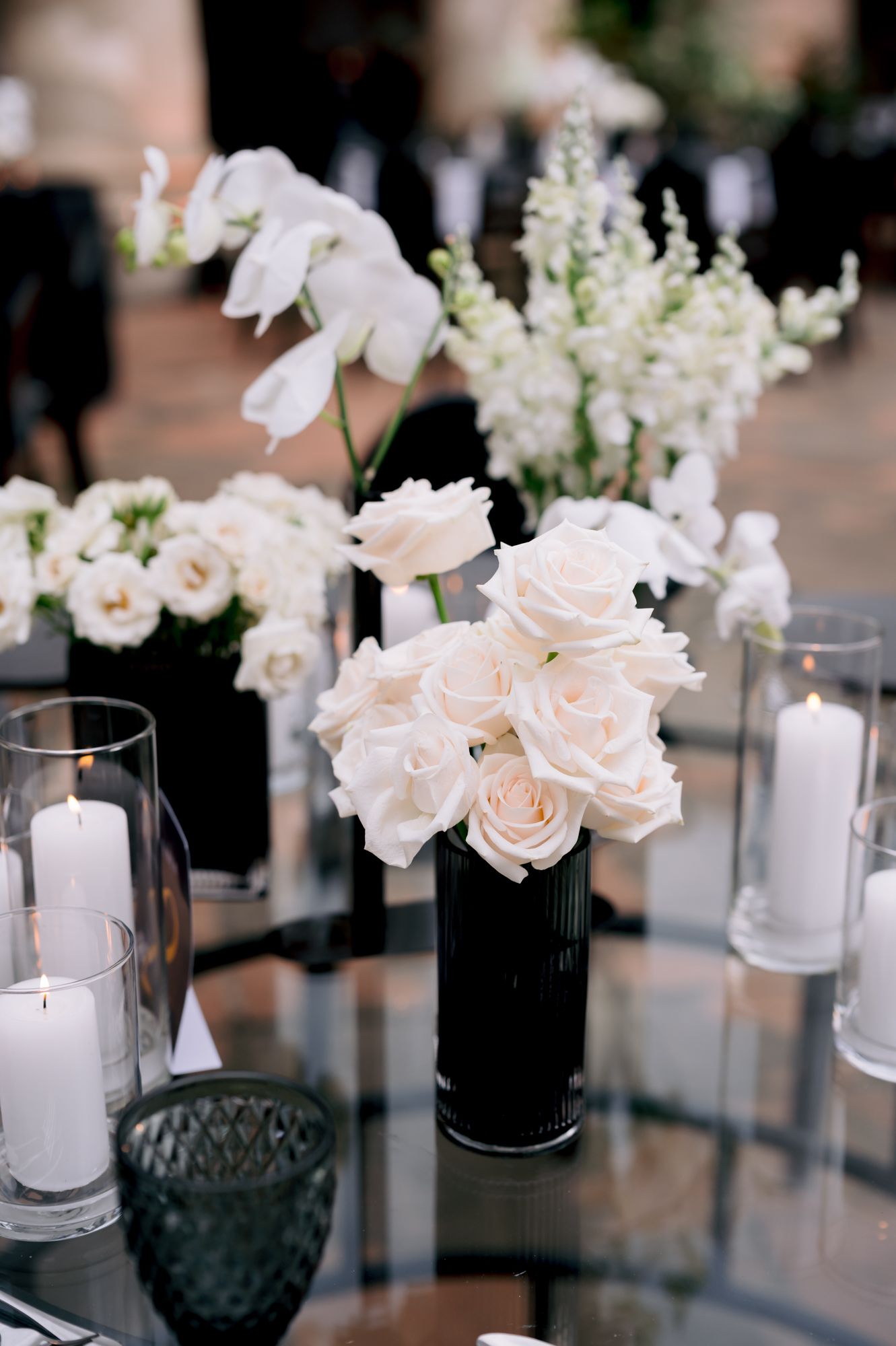 White roses and orchids in black vases with glowing candles on mirrored wedding table.