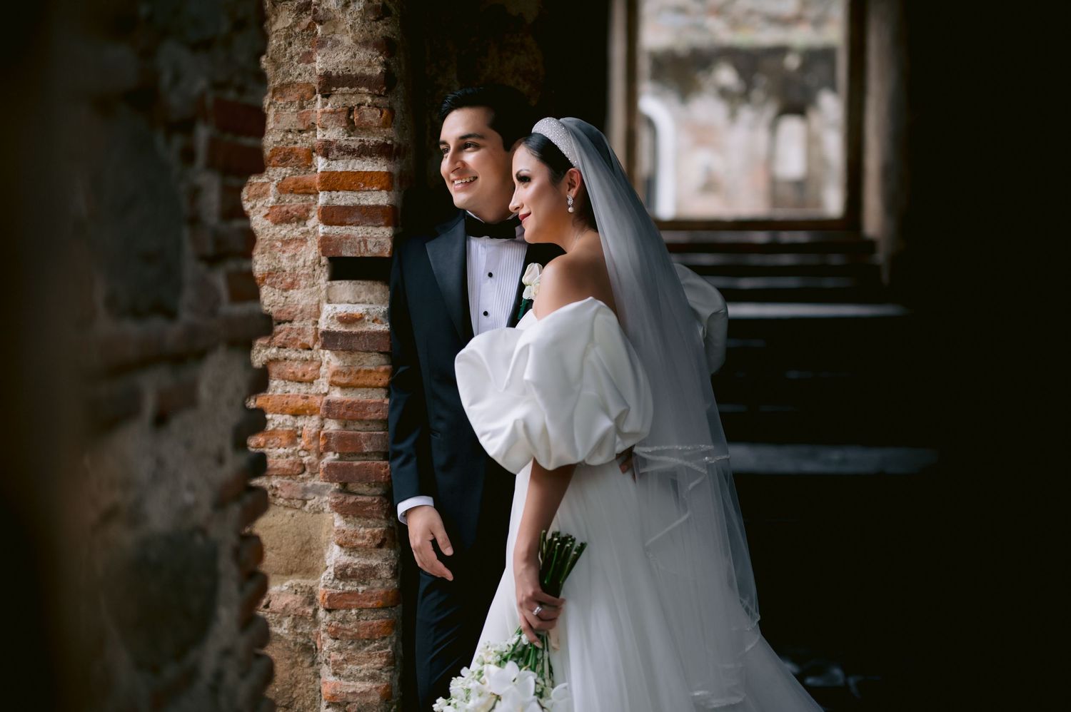 Couple in wedding attire pose together by a rustic brick archway with natural lighting.