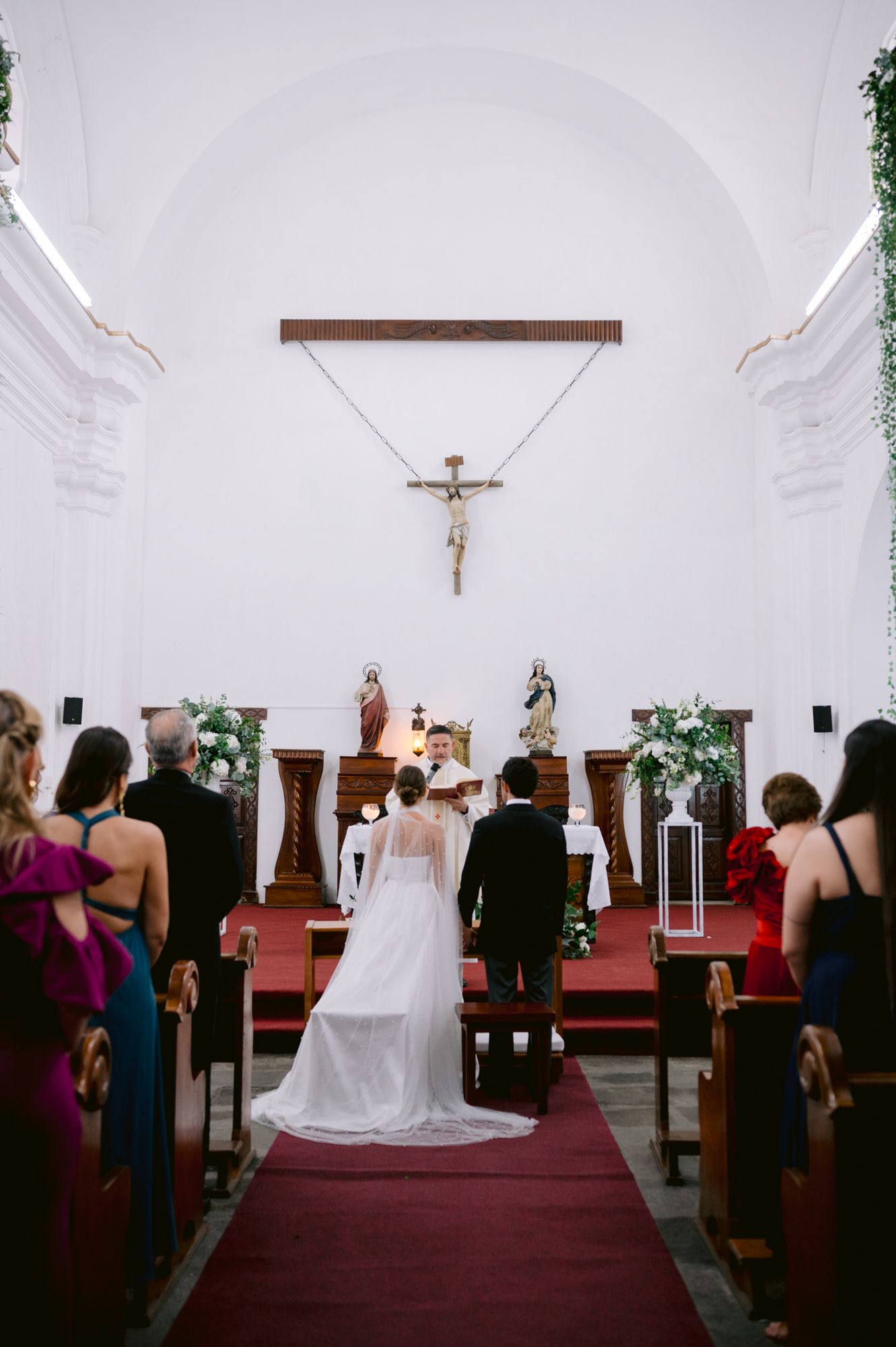 Wedding ceremony in progress inside white church with red carpet aisle and floral decorations.