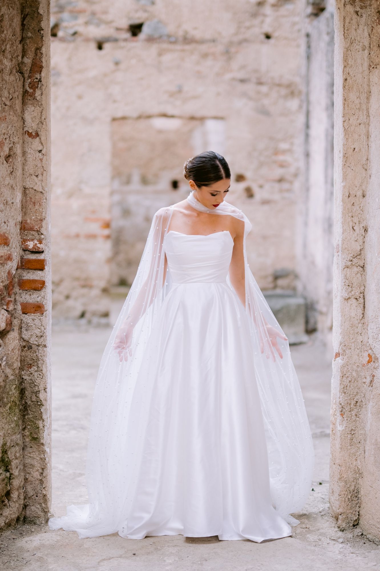 Bride in classic strapless gown with flowing cape stands in ancient stone doorway.