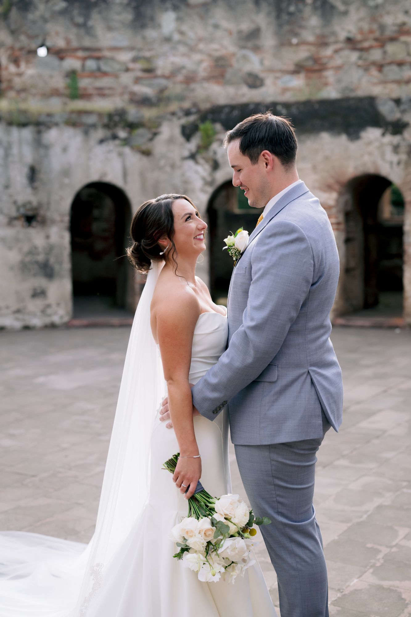 Elegant wedding couple shares an intimate moment against rustic stone walls of historic venue.