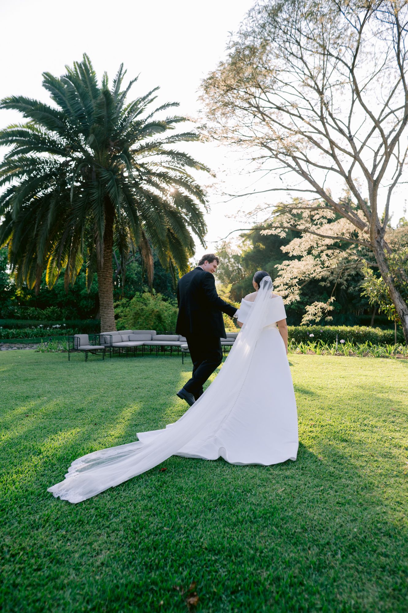 Wedding couple walks through a garden with palm trees and lush greenery as the dress train flows behind.