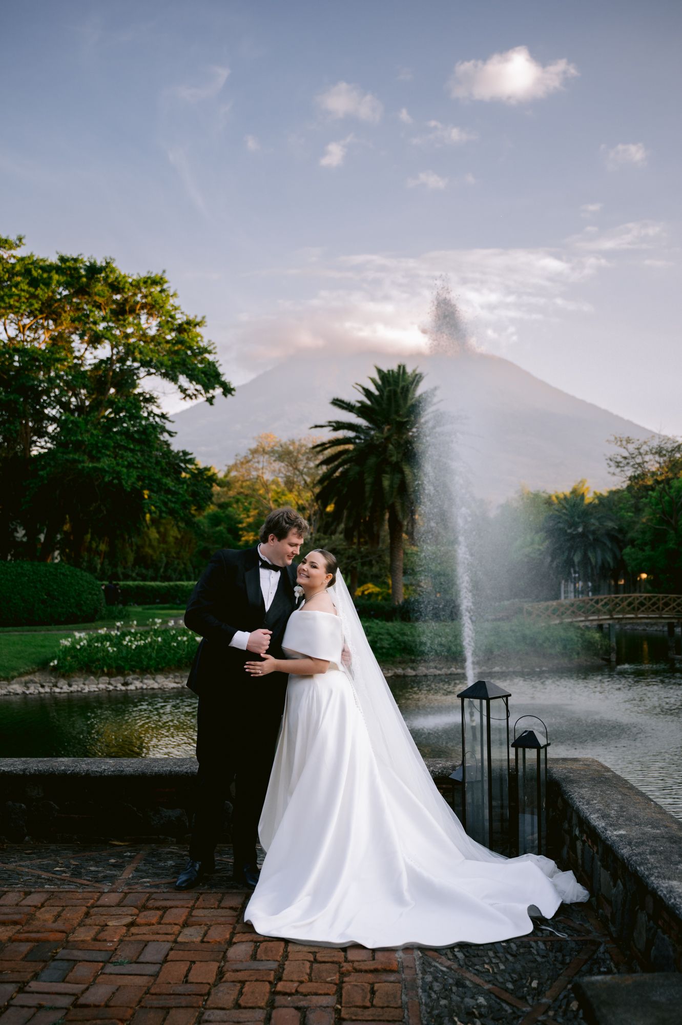Wedding portrait with couple embracing near fountain against mountain landscape at dusk.