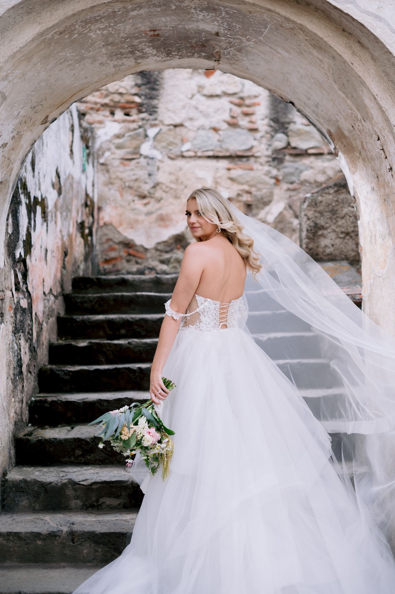 Back view of bride in corset wedding dress with flowing train on historic stone steps.