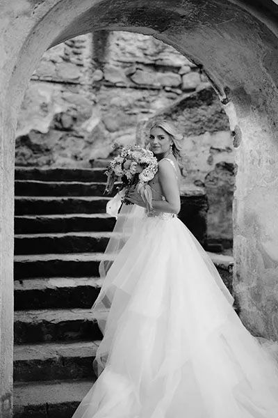 Black and white bridal portrait of person in flowing tulle wedding gown holding bouquet on stone castle steps.