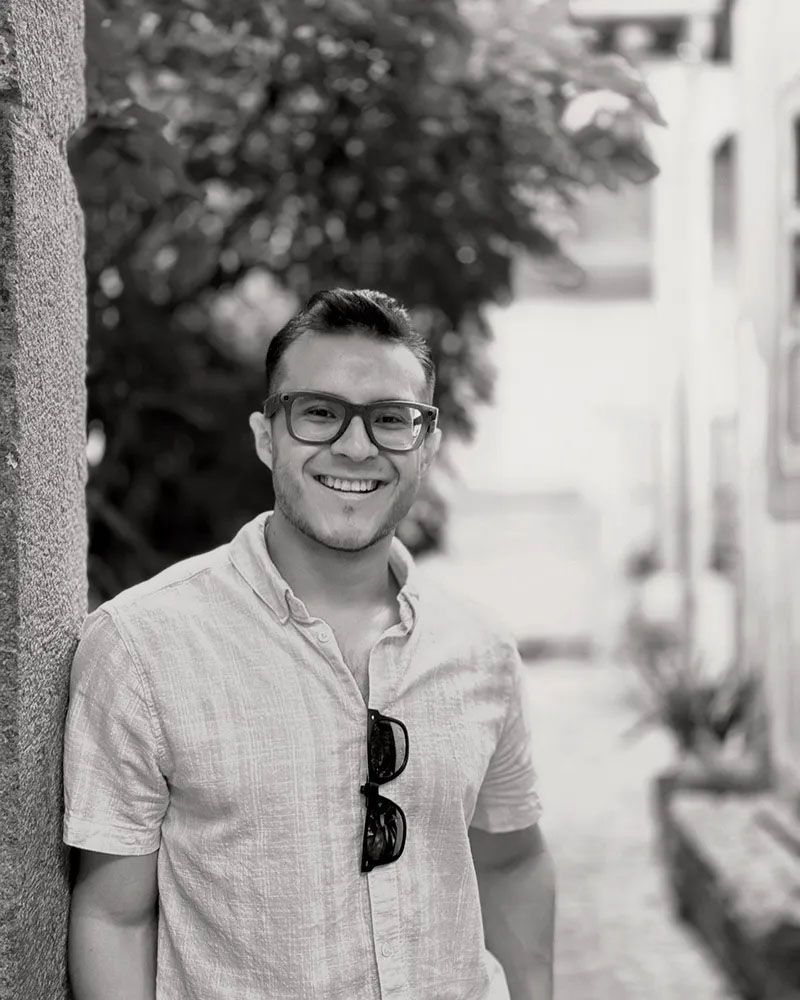 A black and white portrait of someone wearing glasses and a light colored button-down shirt, with a building and trees in background.