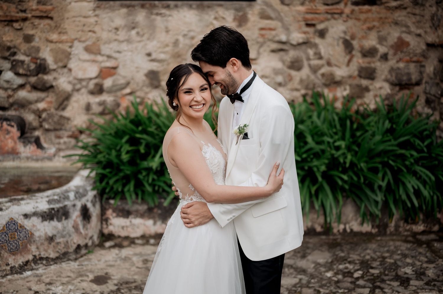 Romantic wedding portrait of couple embracing by stone wall and decorative plants.