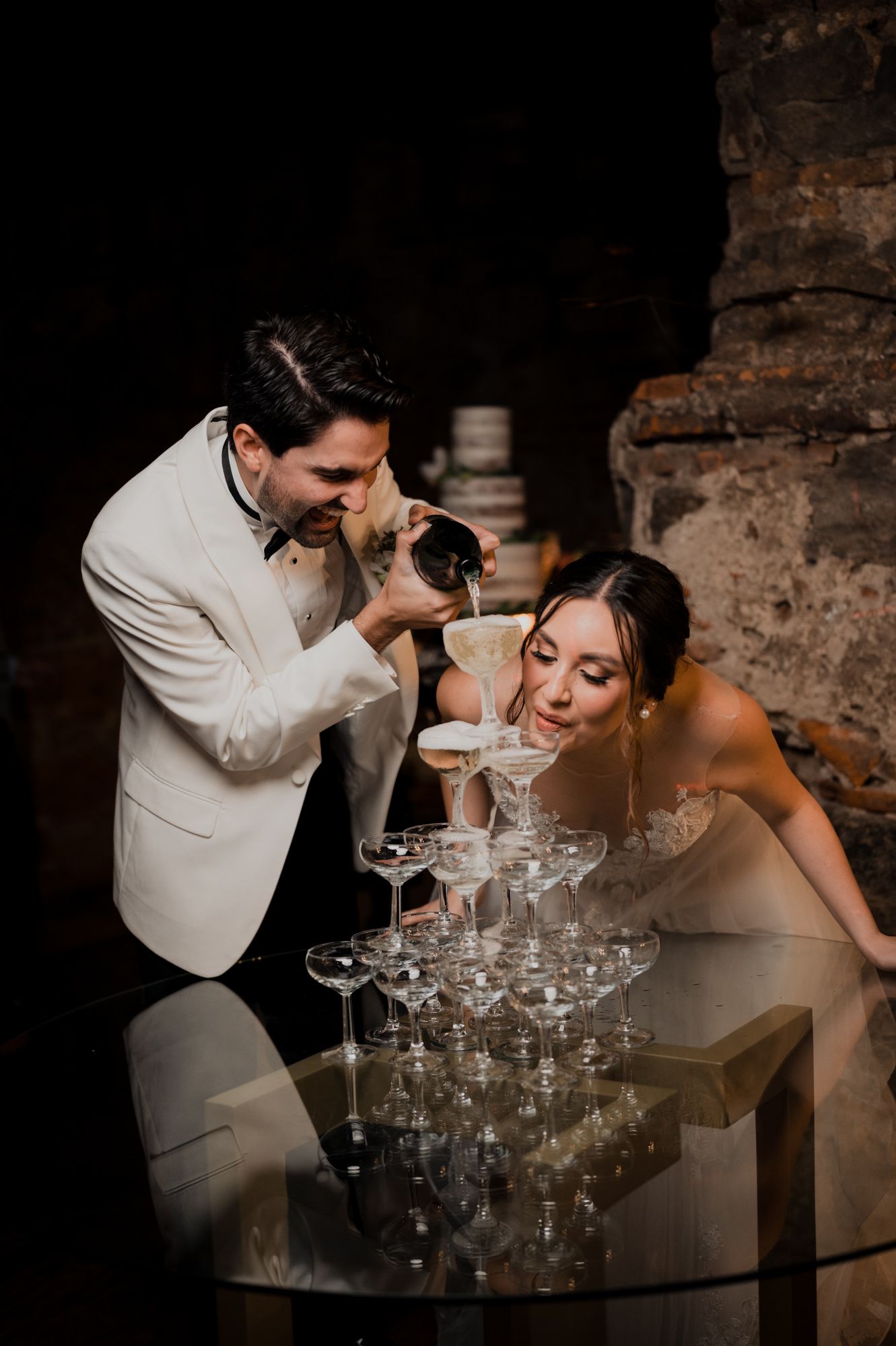 Wedding couple playfully pouring champagne into glass tower during reception celebration.