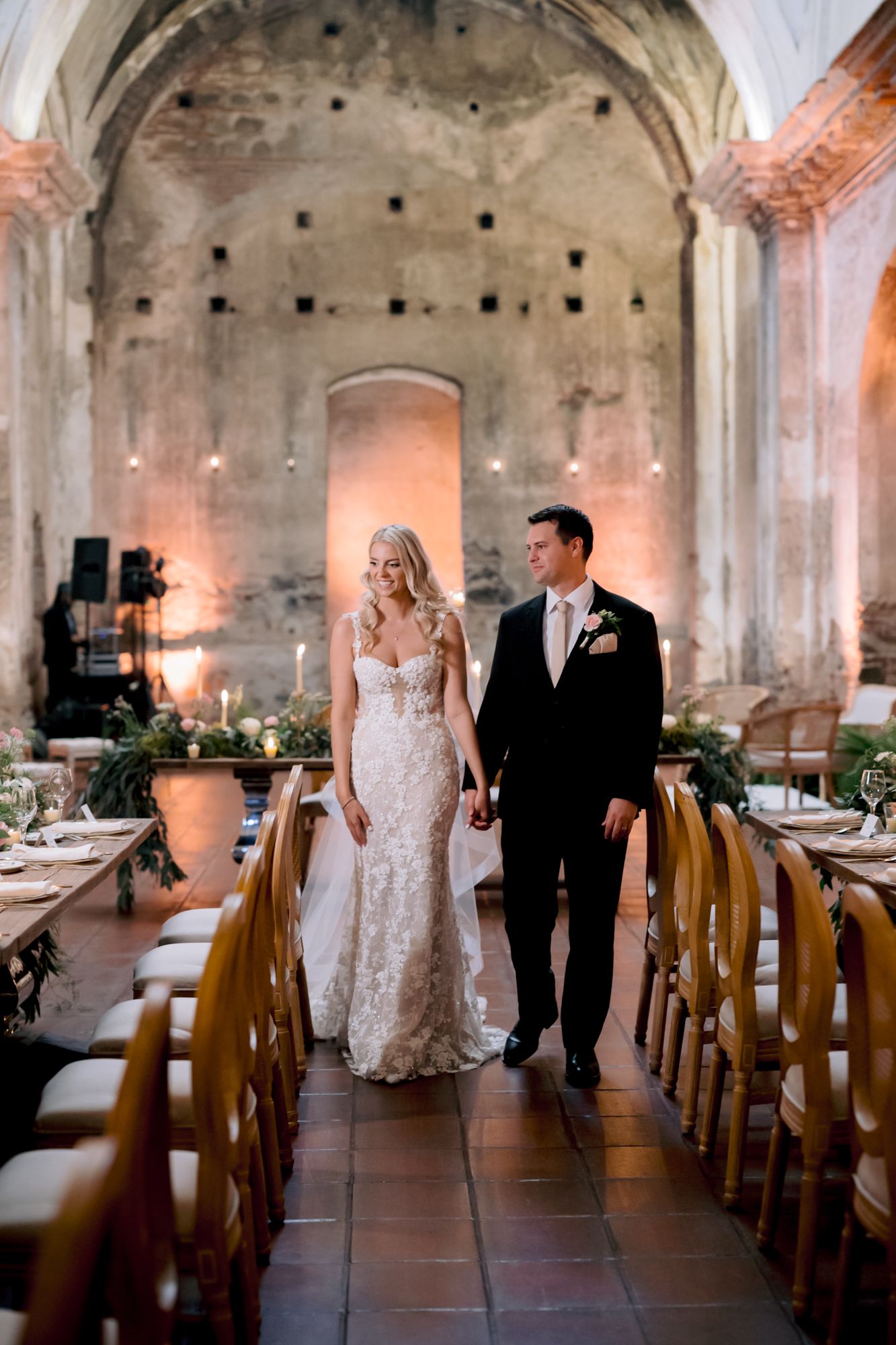 A couple walks through an elegant rustic venue with stone walls and wooden chairs set for a wedding reception.