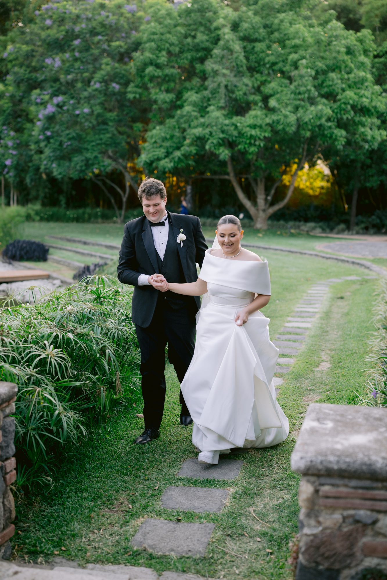Bride and groom walking along garden path surrounded by lush greenery in formal wedding attire.