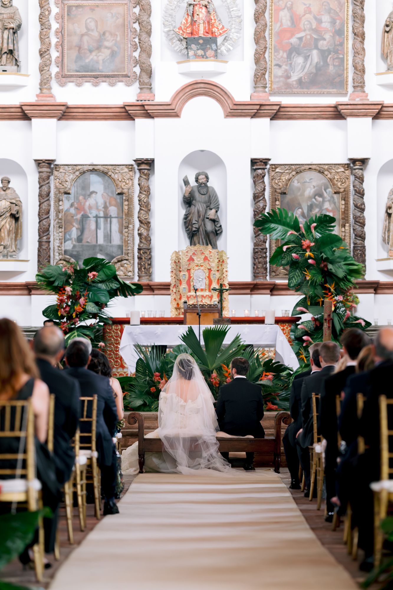 Wedding ceremony in a historic church with religious artwork and tropical greenery decorations.