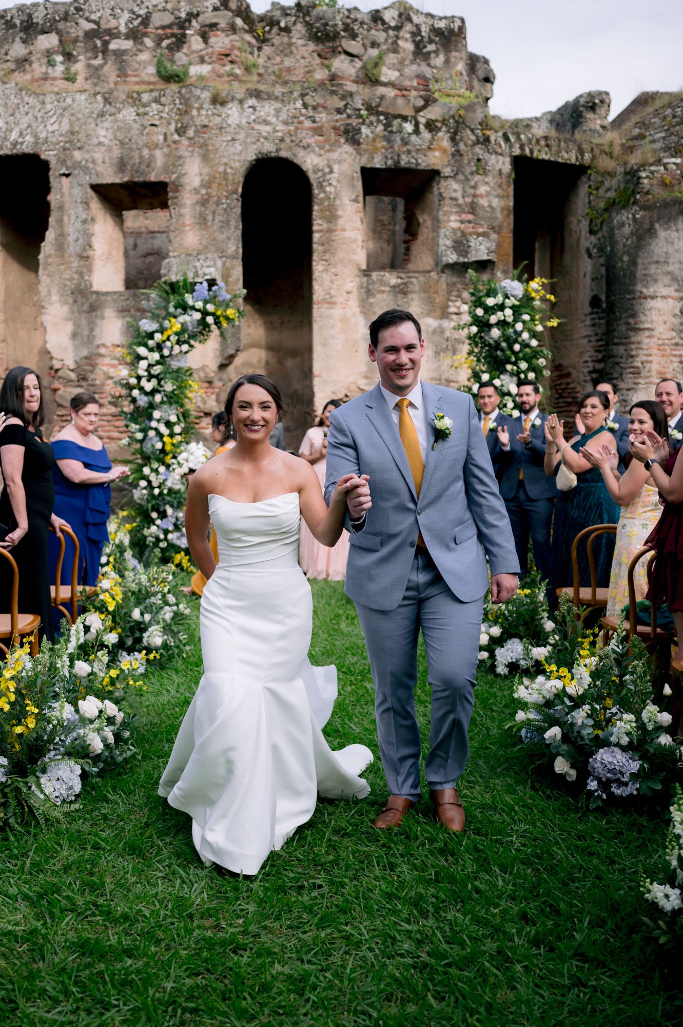 Newlyweds celebrate on flower-lined aisle against backdrop of historic stone ruins.