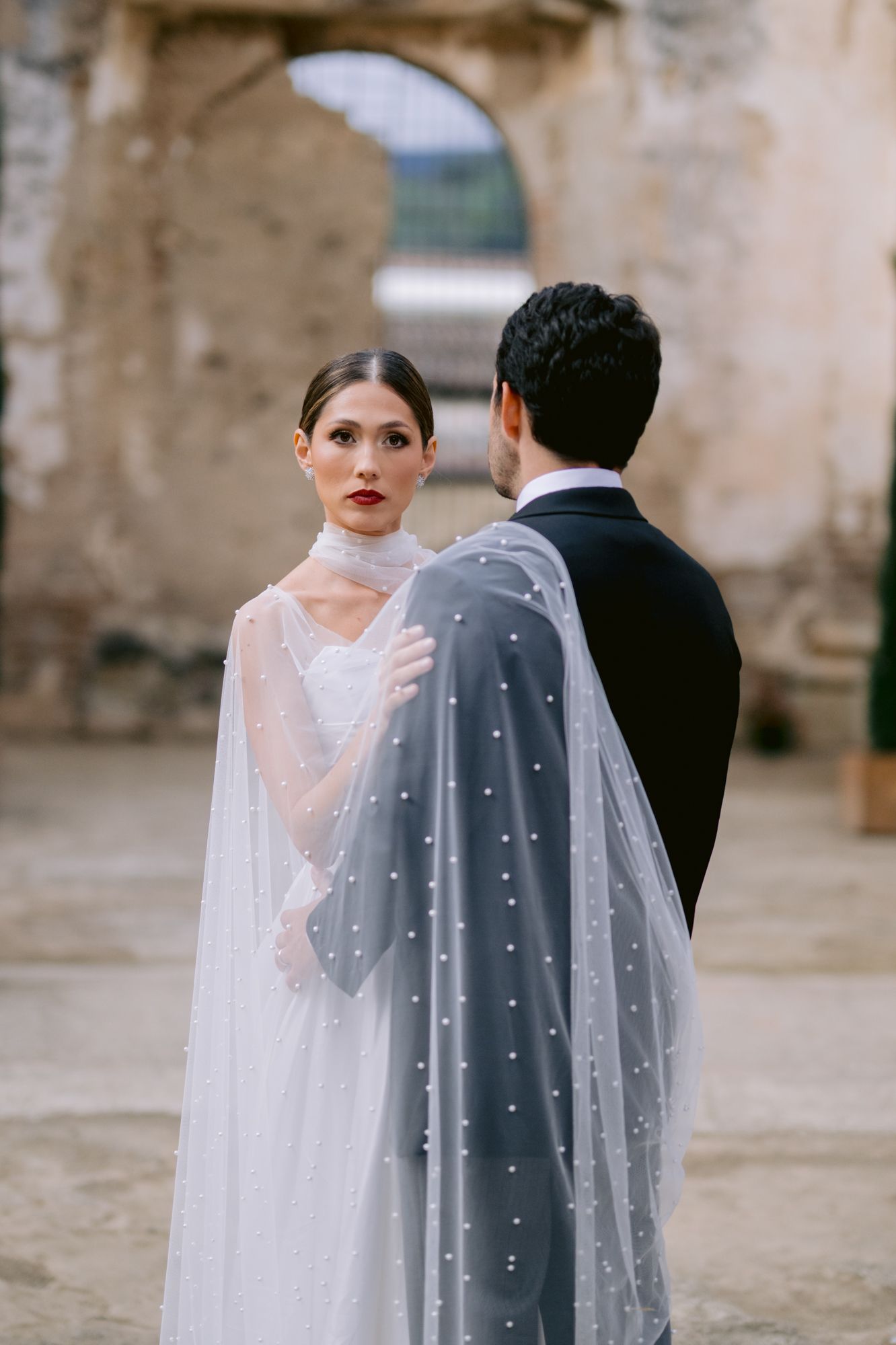 Intimate moment between couple with flowing dotted veil against ancient stone walls.